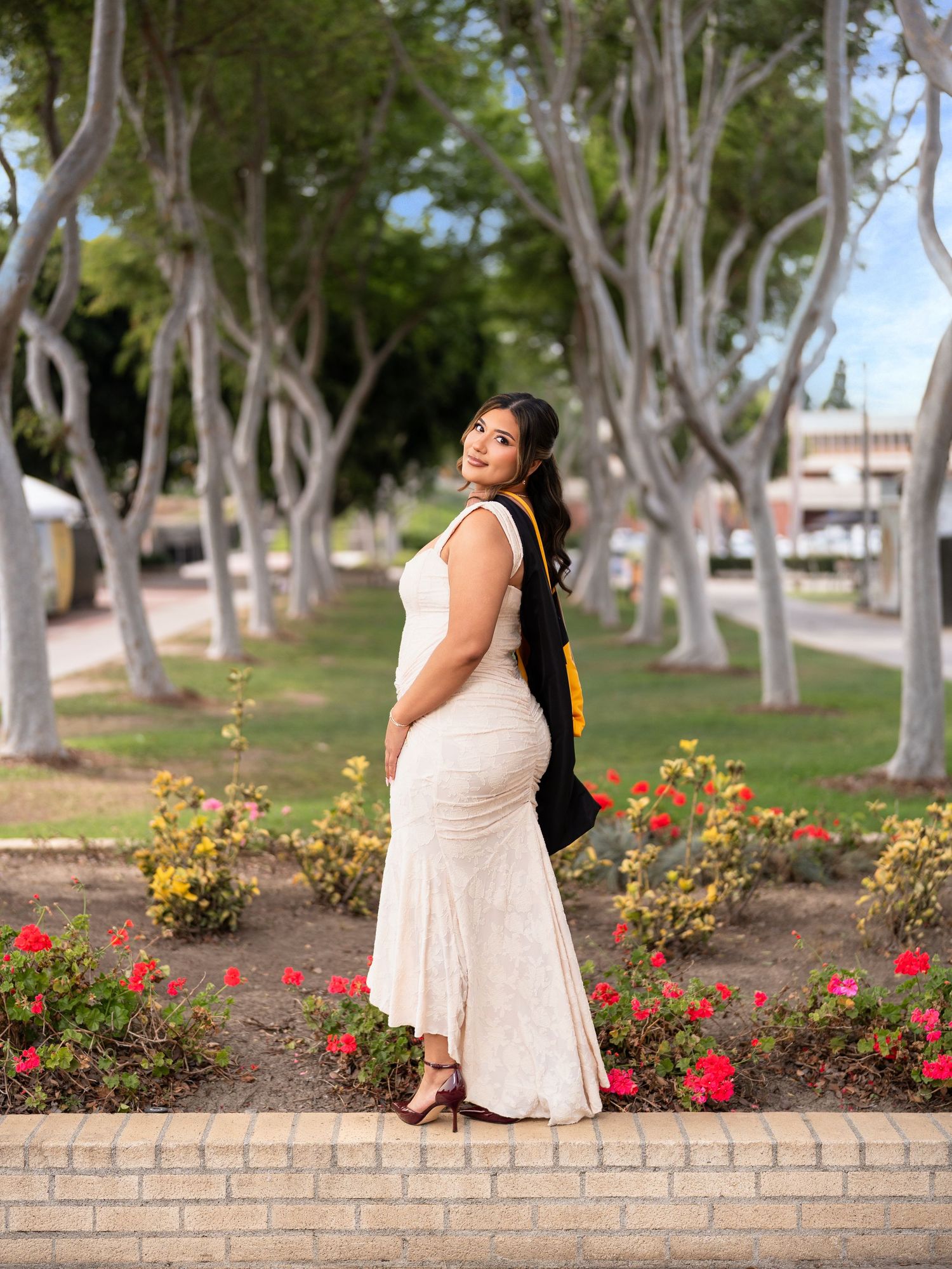 Woman in white dress and graduation regalia poses in a tree-lined garden with red flowers.