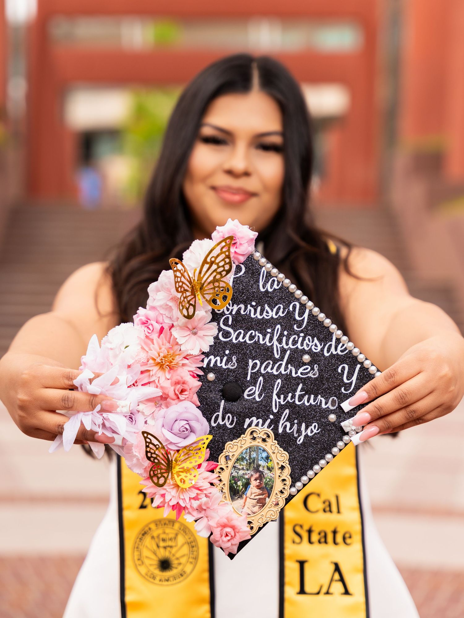A graduation cap decorated with pink flowers, butterflies and text on a black background displays Cal State LA pride.