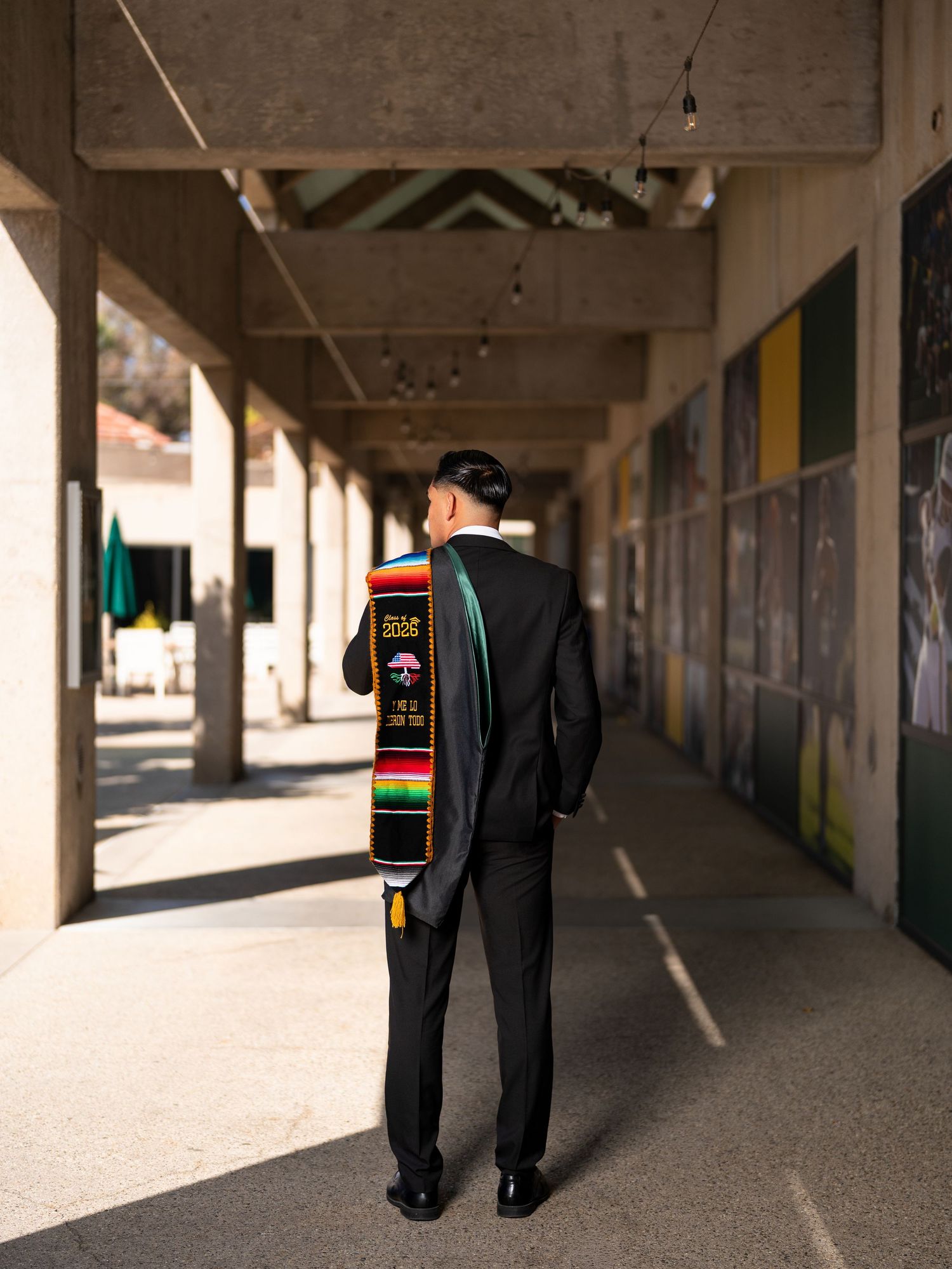 Graduate in black suit with colorful cultural stole stands in sunlit campus corridor, viewed from behind.