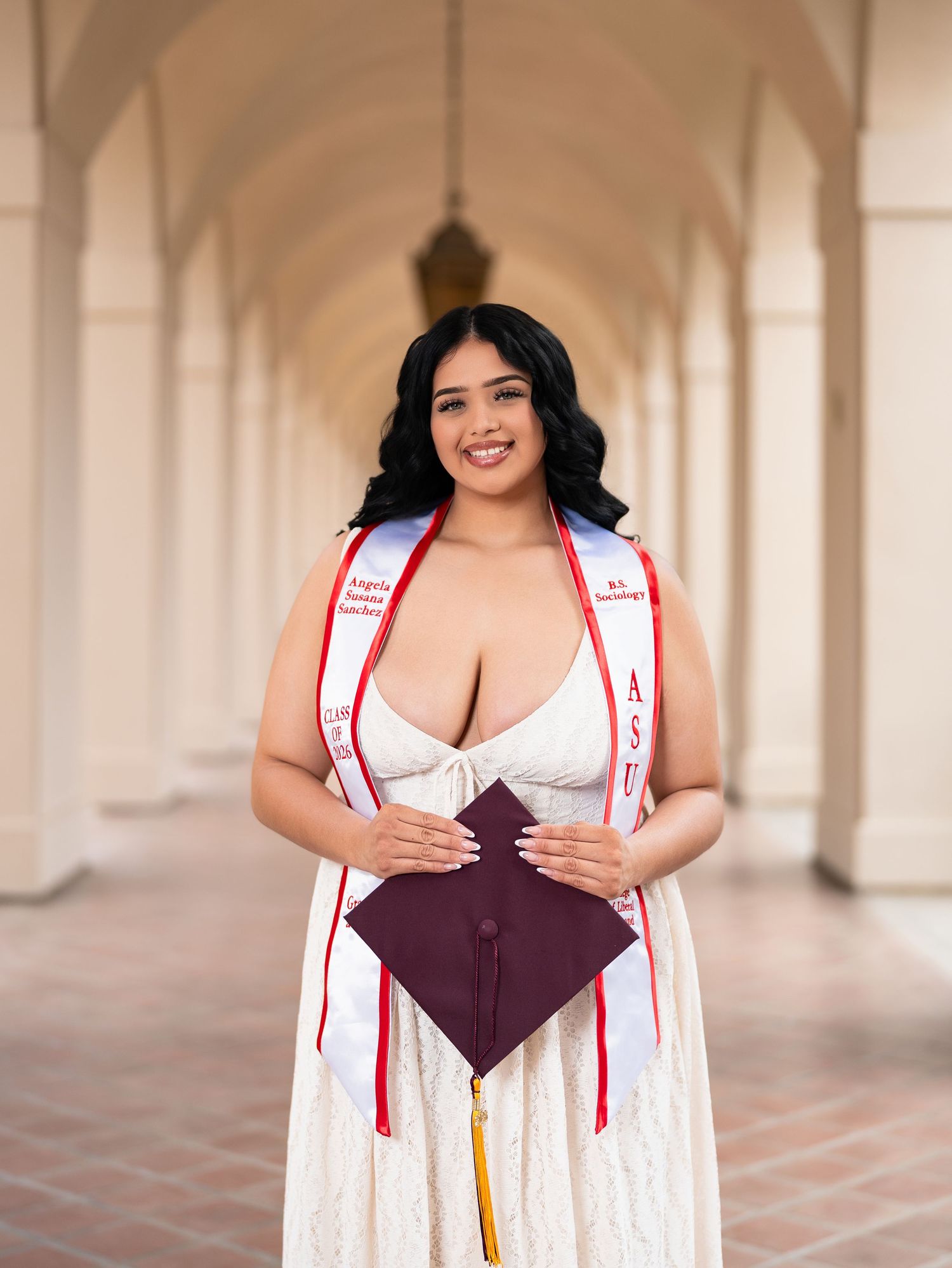Young woman in white lace dress holding maroon graduation cap, wearing ASU sash, posing under arched colonnade.
