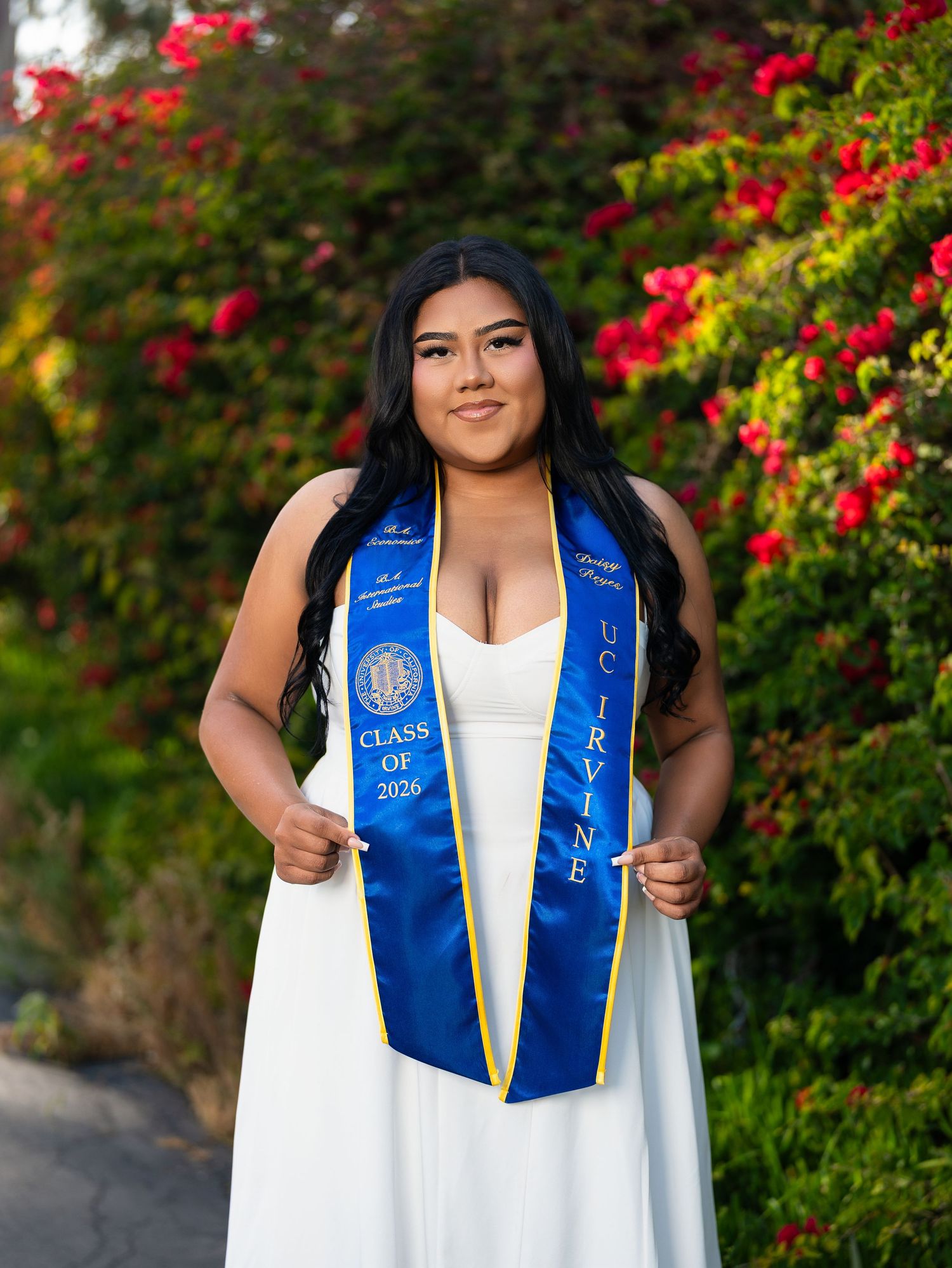 UC Irvine graduate in white dress and blue Class of 2026 graduation stole posing outdoors with red flowers.