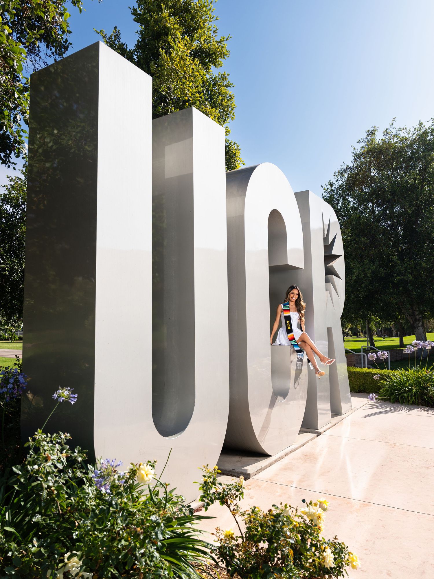 Woman sitting inside large UCR letter sculpture on sunny campus grounds with lush green trees.