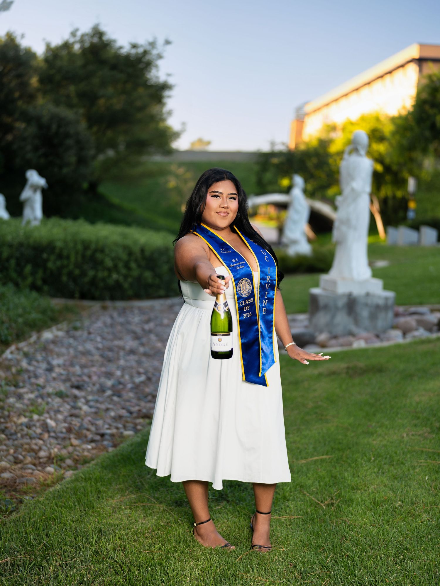 Graduate in white dress and blue sash holds champagne bottle in garden with statues.