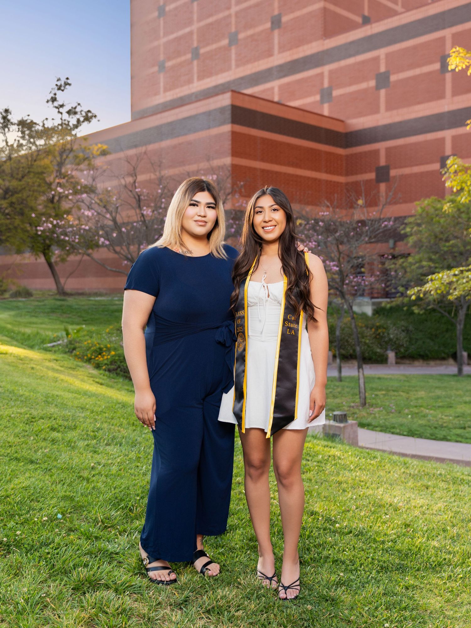 Two people stand together on a grassy slope in front of a brick building on a sunny afternoon.