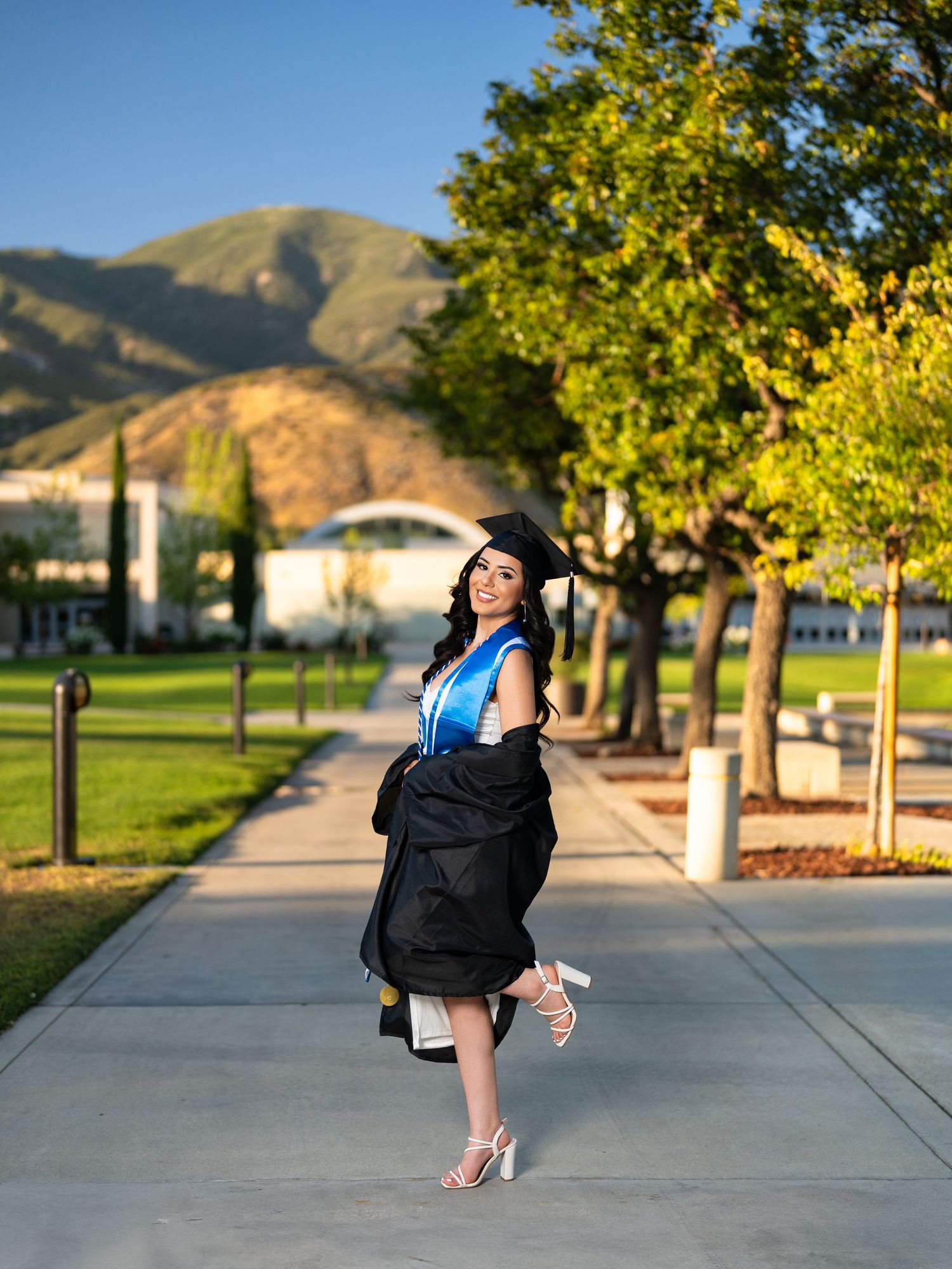 A woman in graduation cap and gown walks on a campus path with green trees and mountains in the background.
