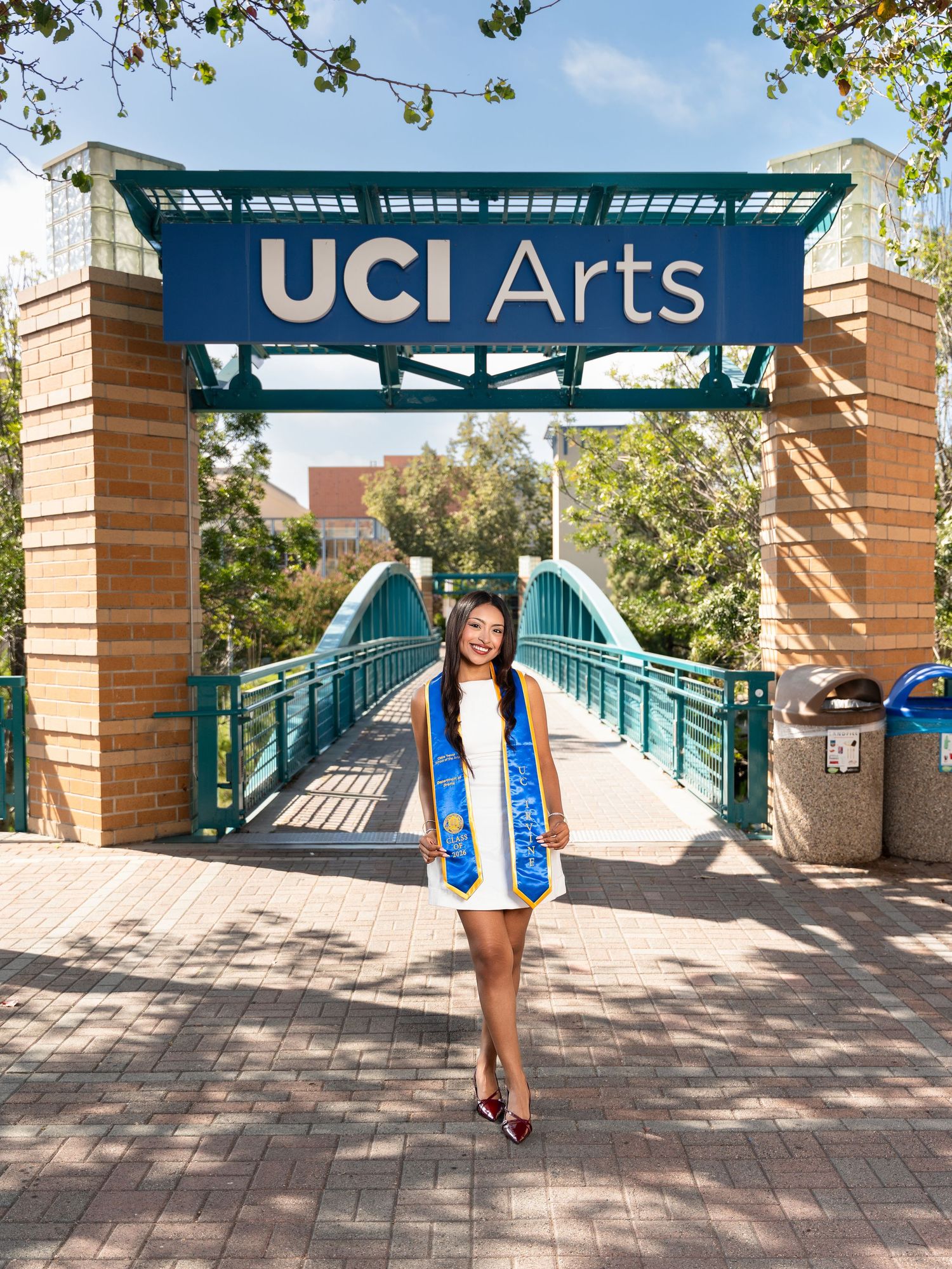 Graduate poses under UCI Arts archway in cap and gown regalia on bridge at UC Irvine campus.