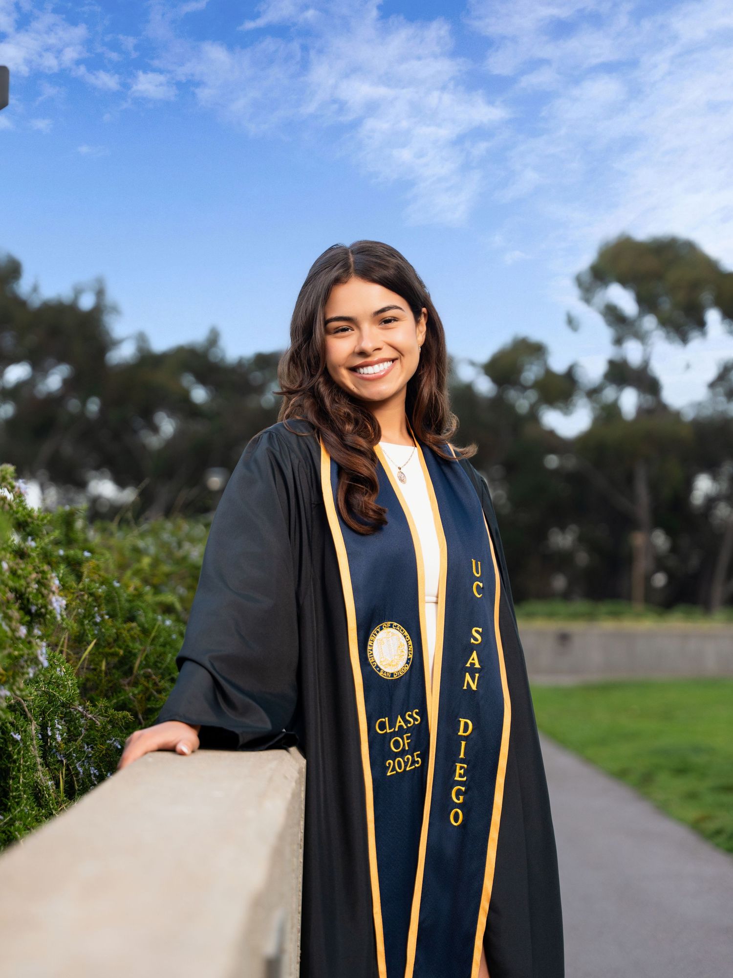 A graduate wearing a dark academic gown with gold sash stands outdoors against a blue sky and trees on a sunny day.