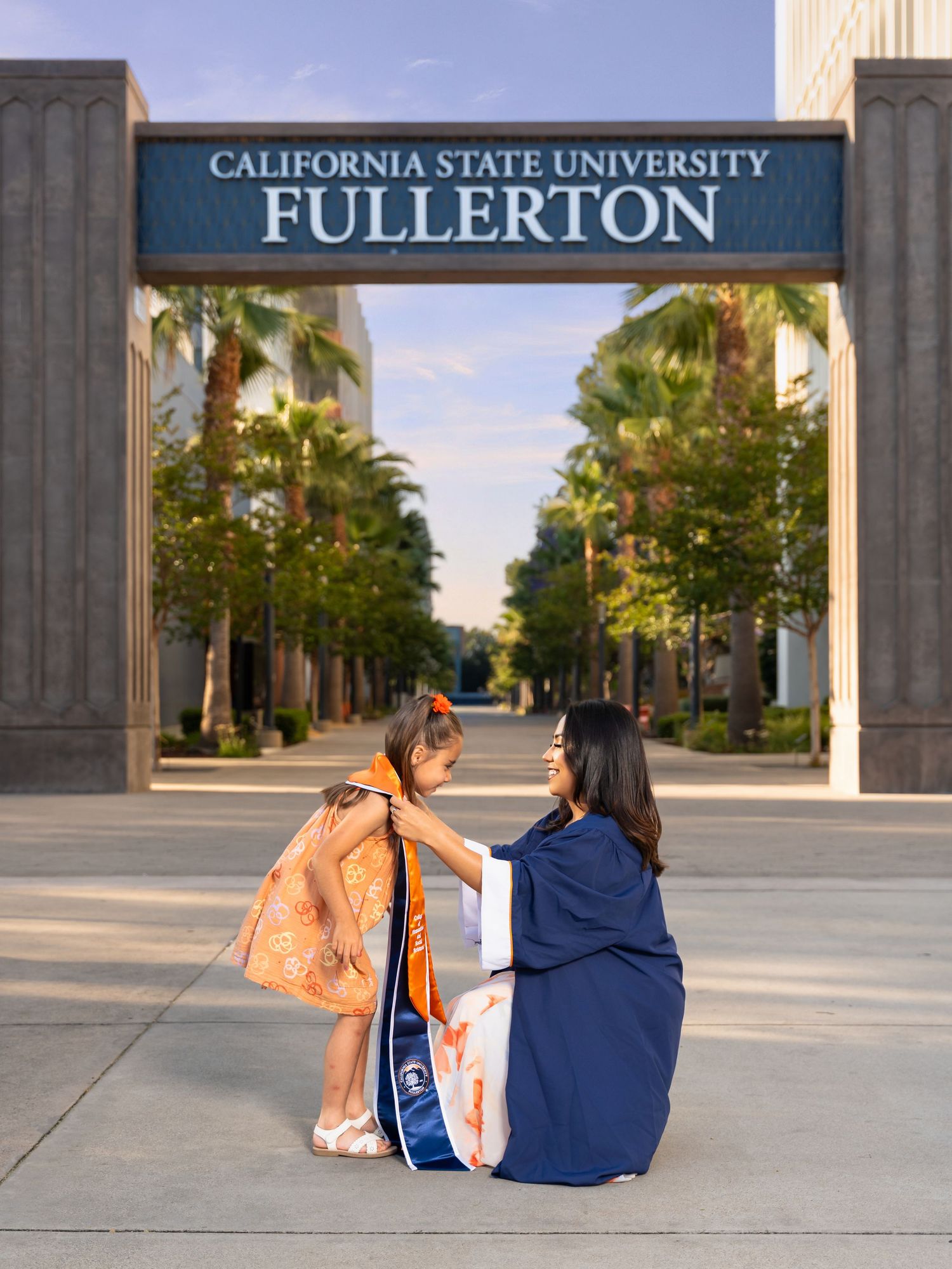 Two students sit on the ground beneath the California State University Fullerton entrance sign on a palm tree-lined path.