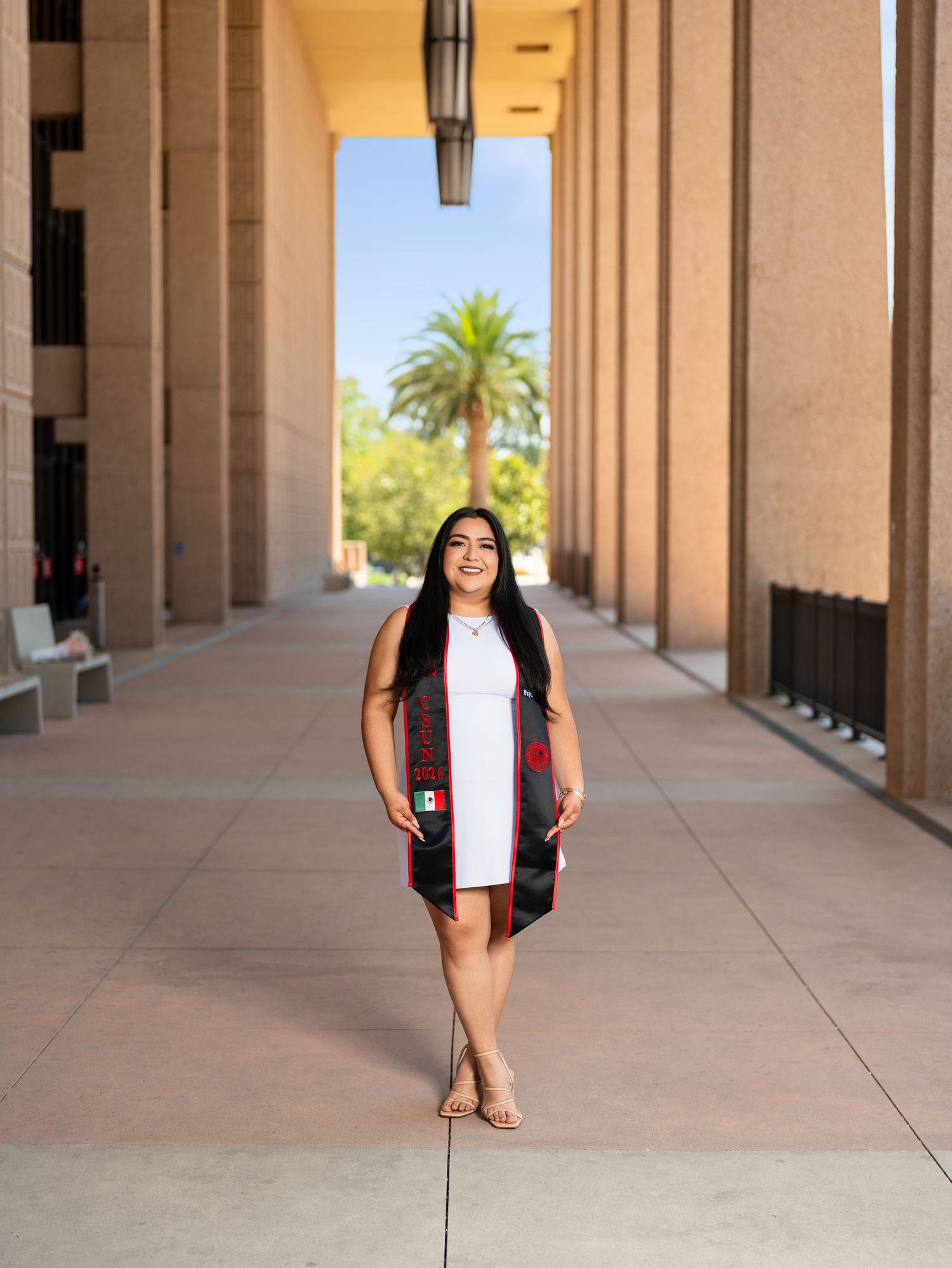 A smiling graduate poses in a white dress with a colorful stole between columns on a sunny campus walkway.