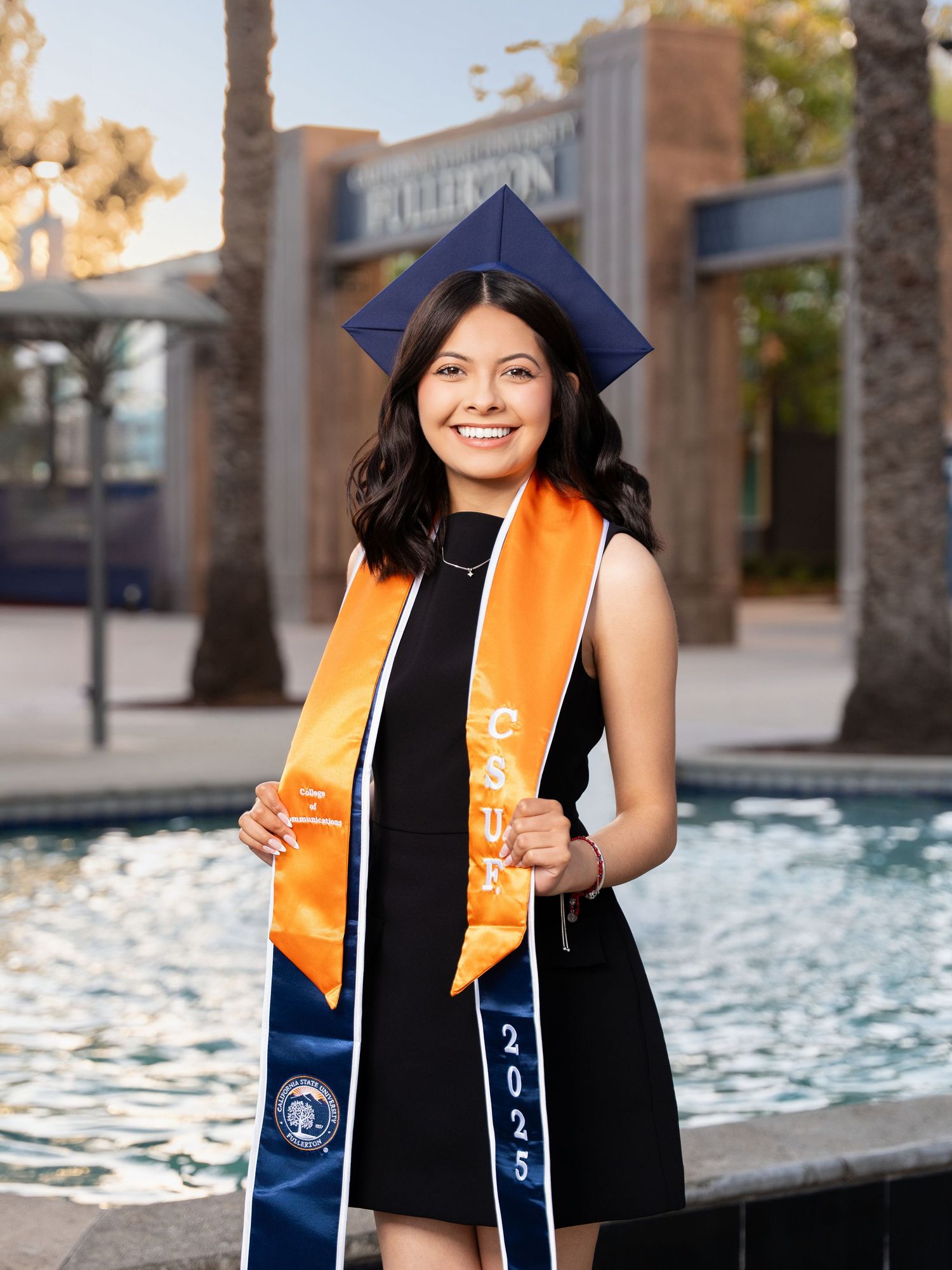 Graduate wearing black cap and orange stole poses for graduation photos by a reflective pool.
