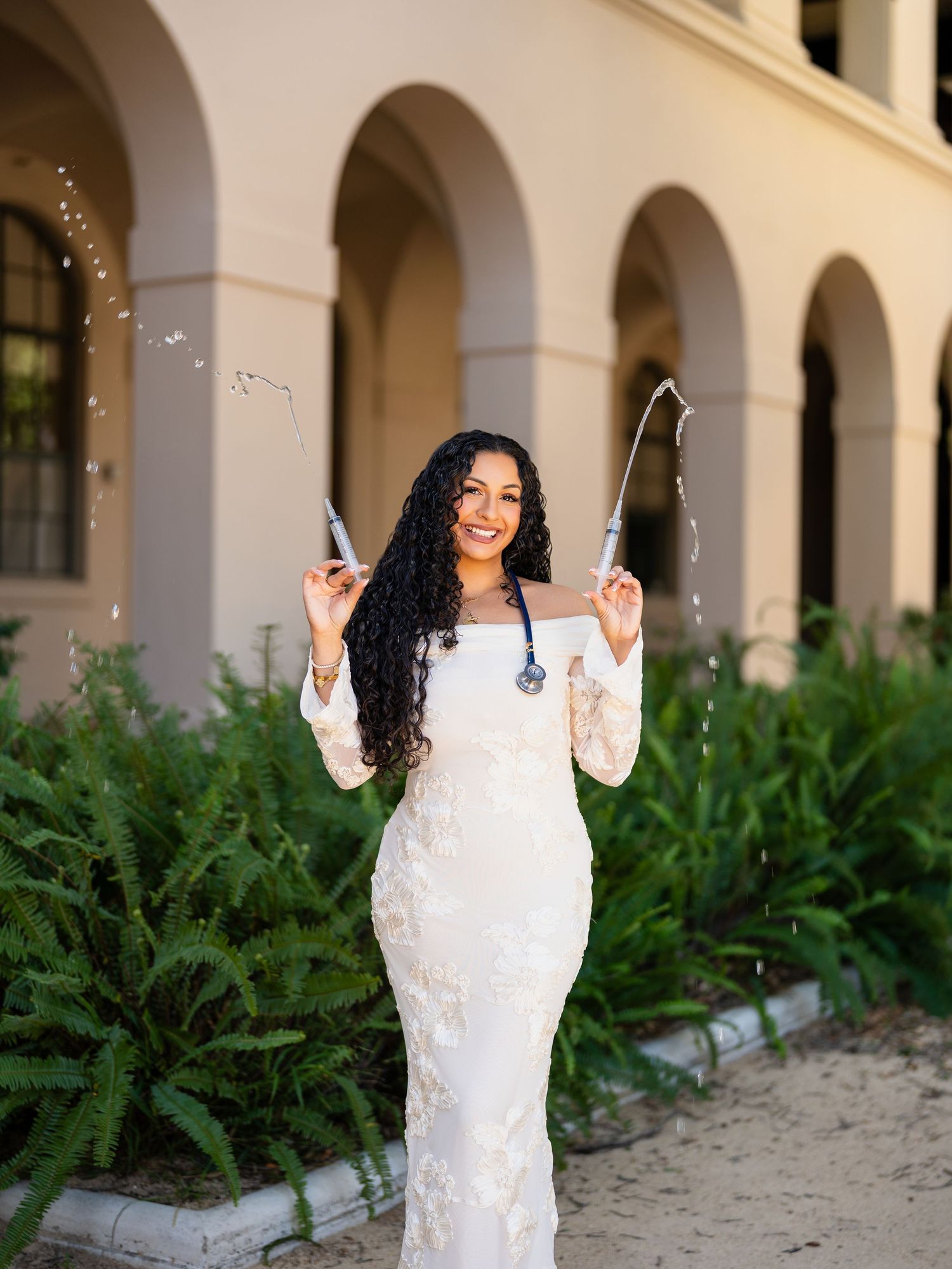 A person in a white lace dress poses gracefully against a backdrop of arched columns and green foliage.