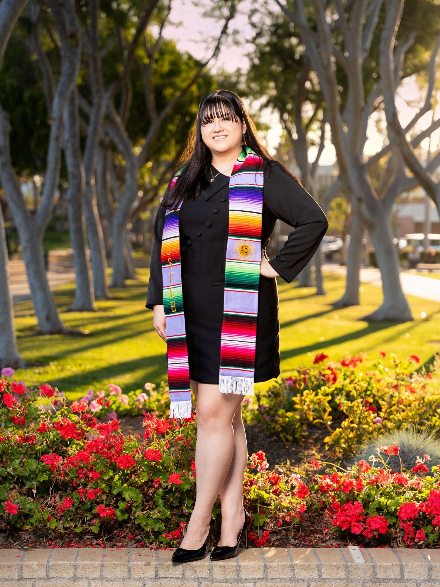 A woman in a black dress and colorful striped scarf poses in front of blooming red flowers and trees at sunset.
