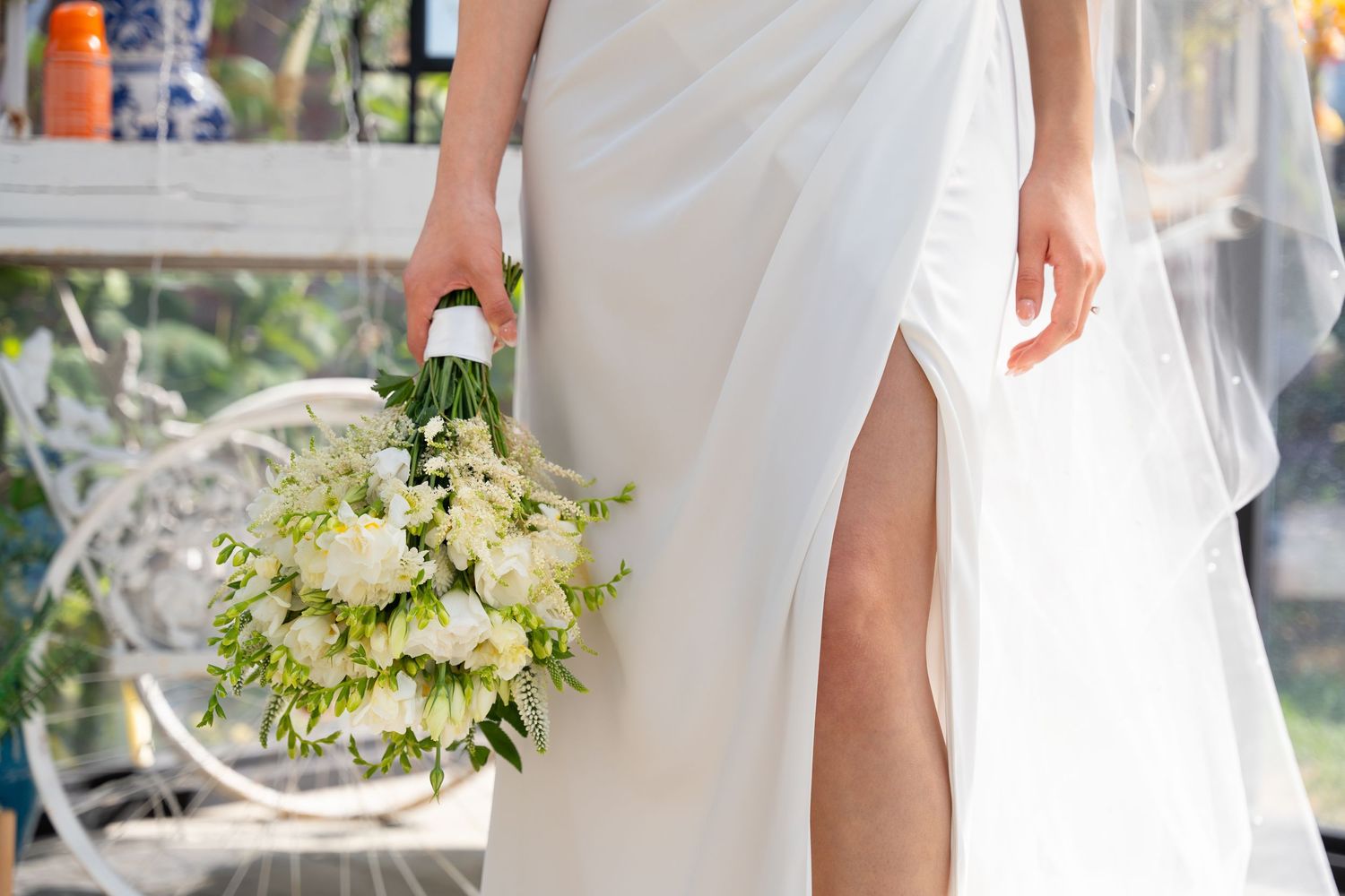 A bride in a flowing white gown holds a delicate bouquet of white and green flowers while showing an elegant leg slit.
