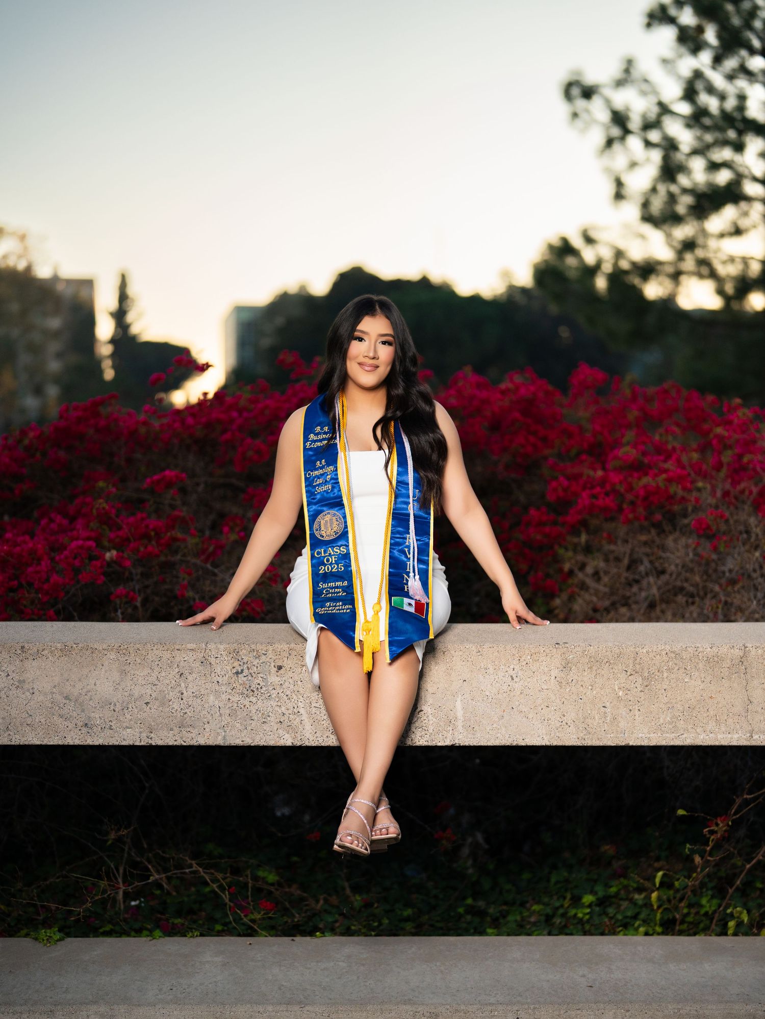 Graduate in blue and gold stole sits on concrete wall with vibrant red flowers blooming in background at sunset.