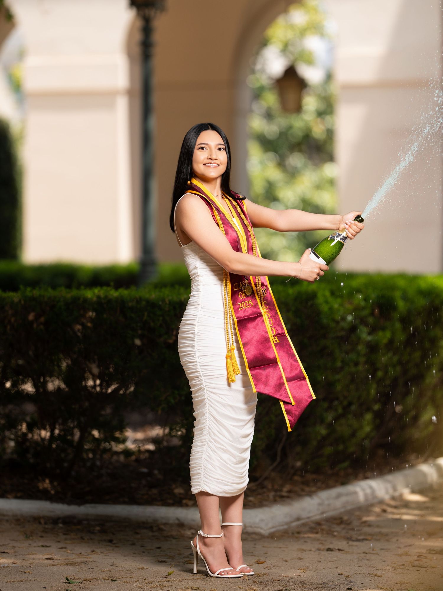 Graduate in white dress and red sash poses outdoors during a photo session at graduation.