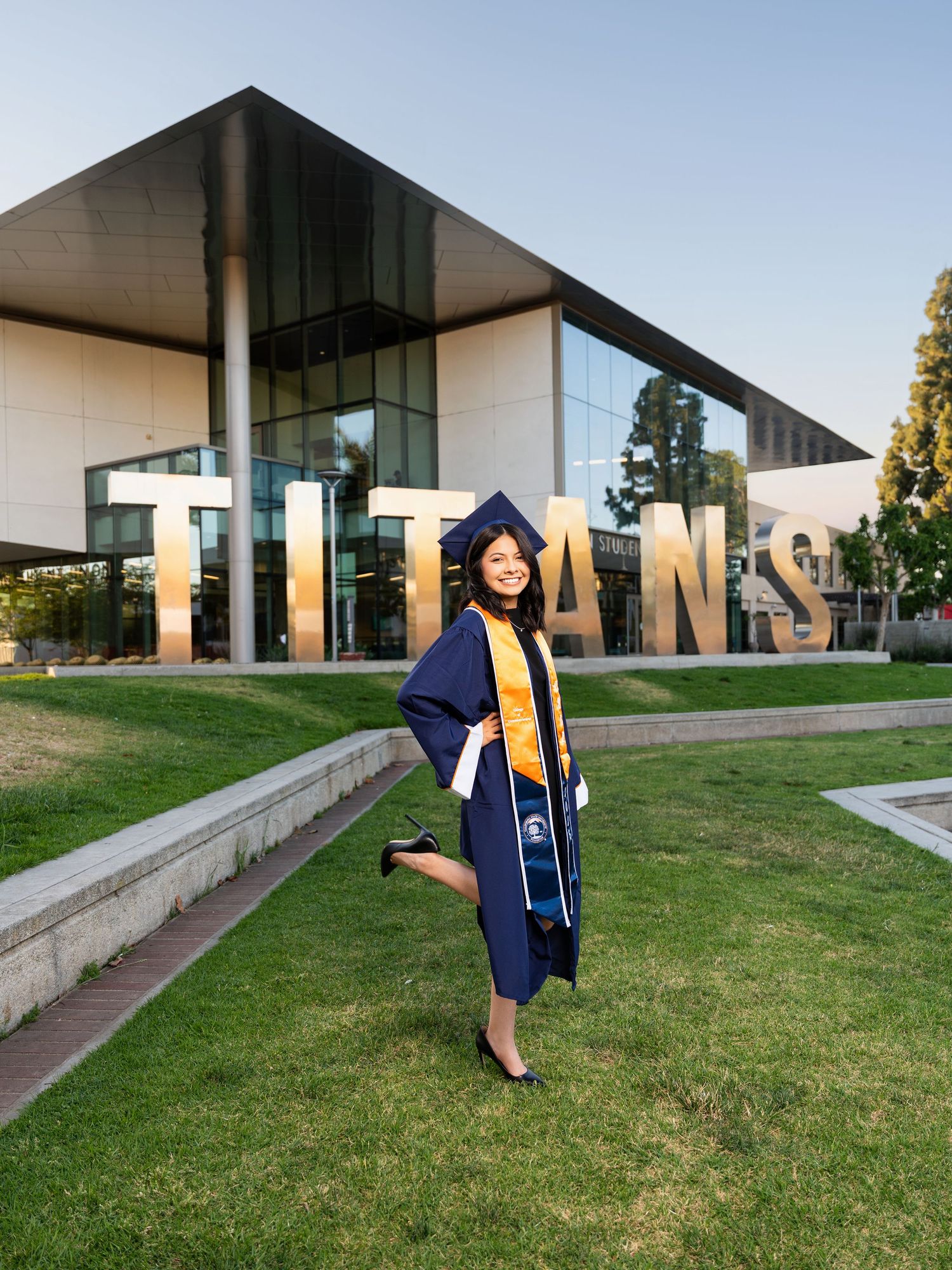 Graduate in navy blue cap and gown poses on grassy lawn in front of modern campus building at sunset.