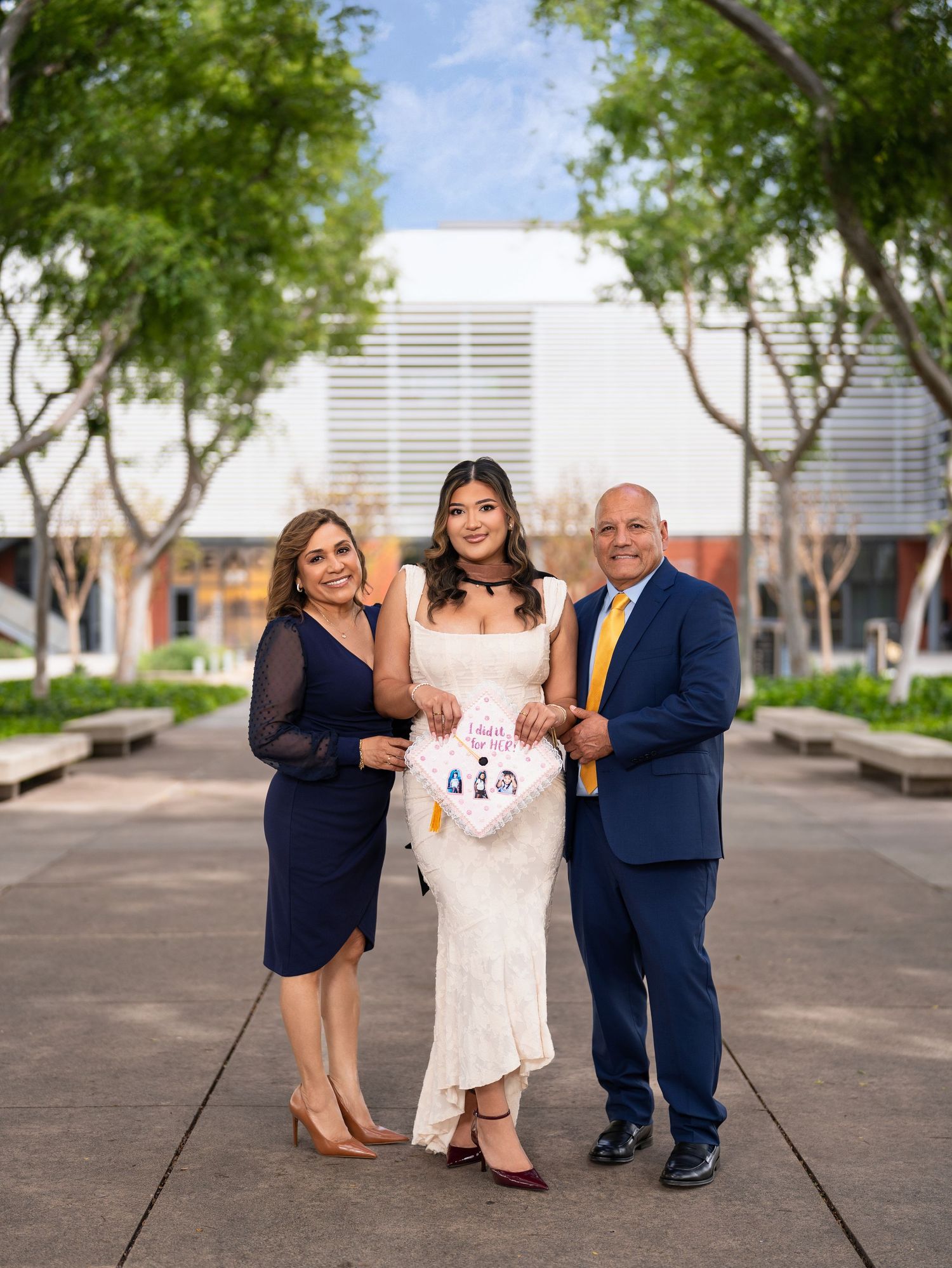 A young woman in a white lace dress holds a stuffed animal flanked by her parents in formal attire outdoors.