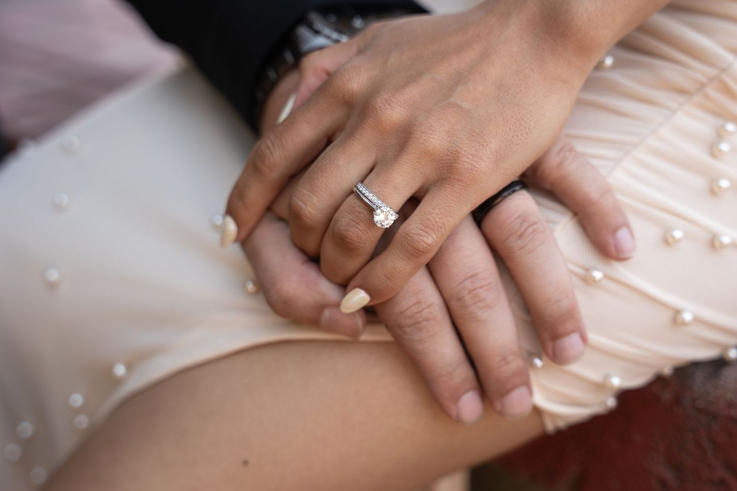 Close-up of hands wearing engagement rings against a light pink dress fabric with delicate beaded details.