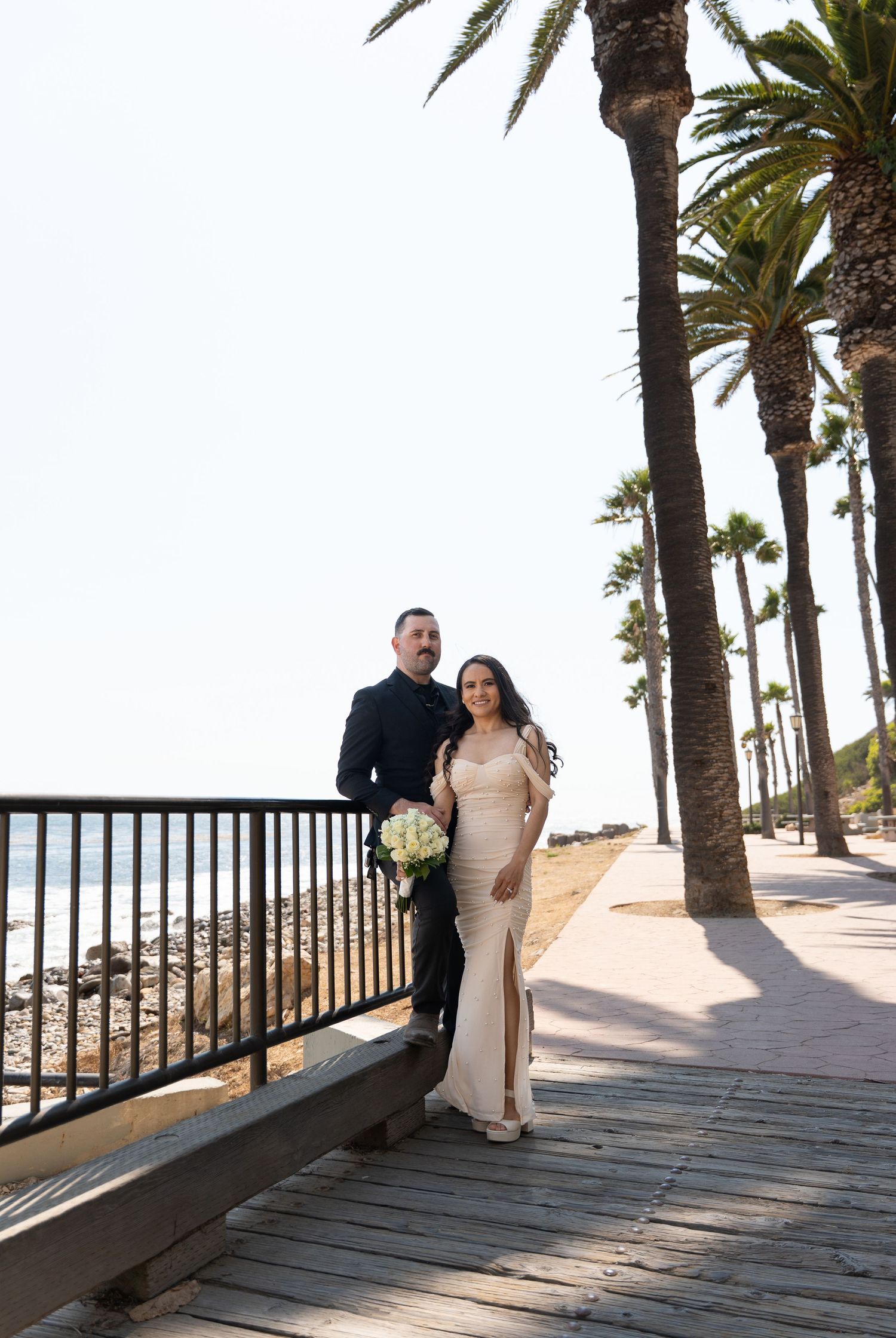 A couple strolls along a palm tree-lined boardwalk near the ocean.