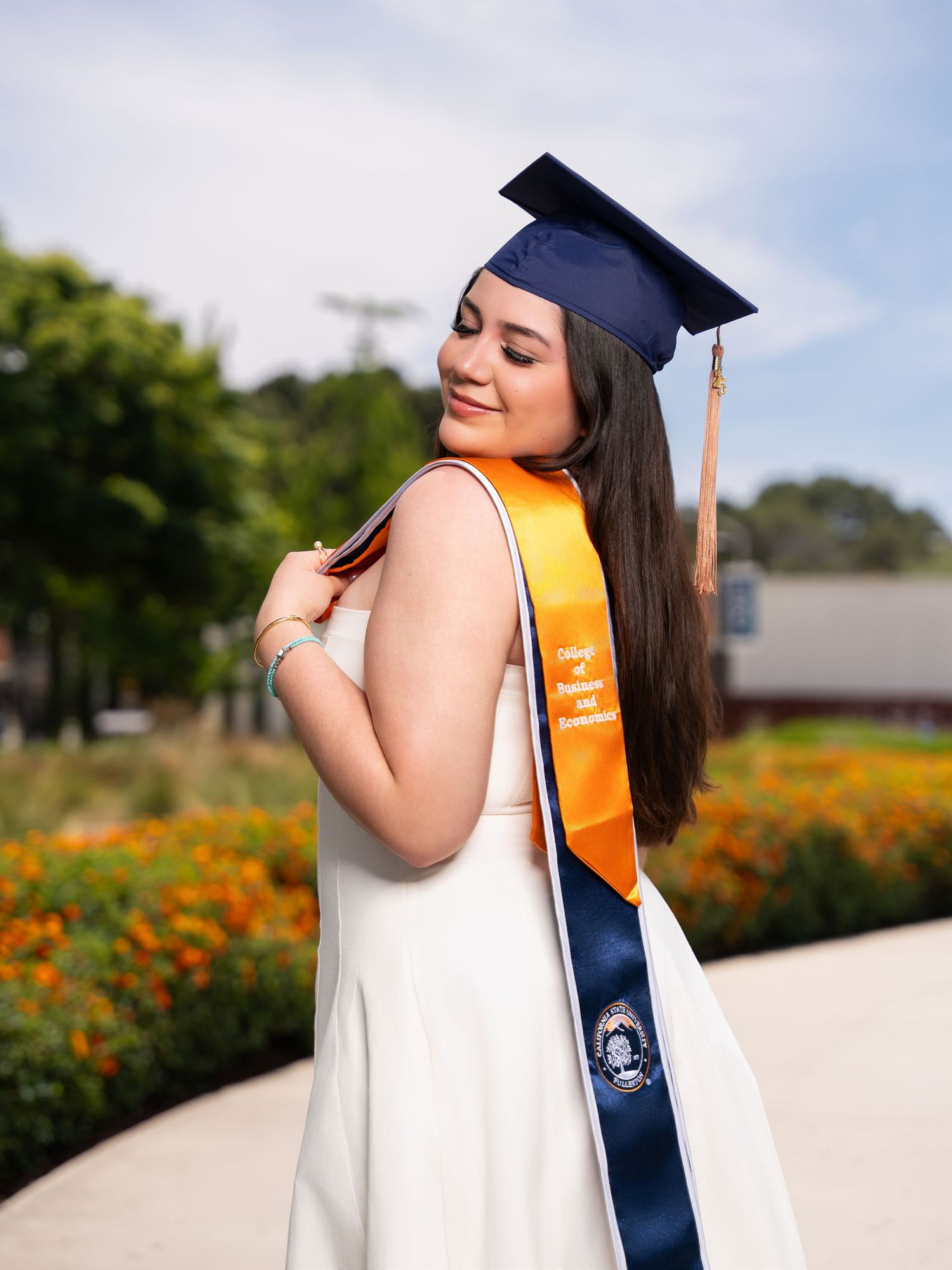Graduate in white dress and navy cap poses with orange sash against outdoor garden setting with flowers.