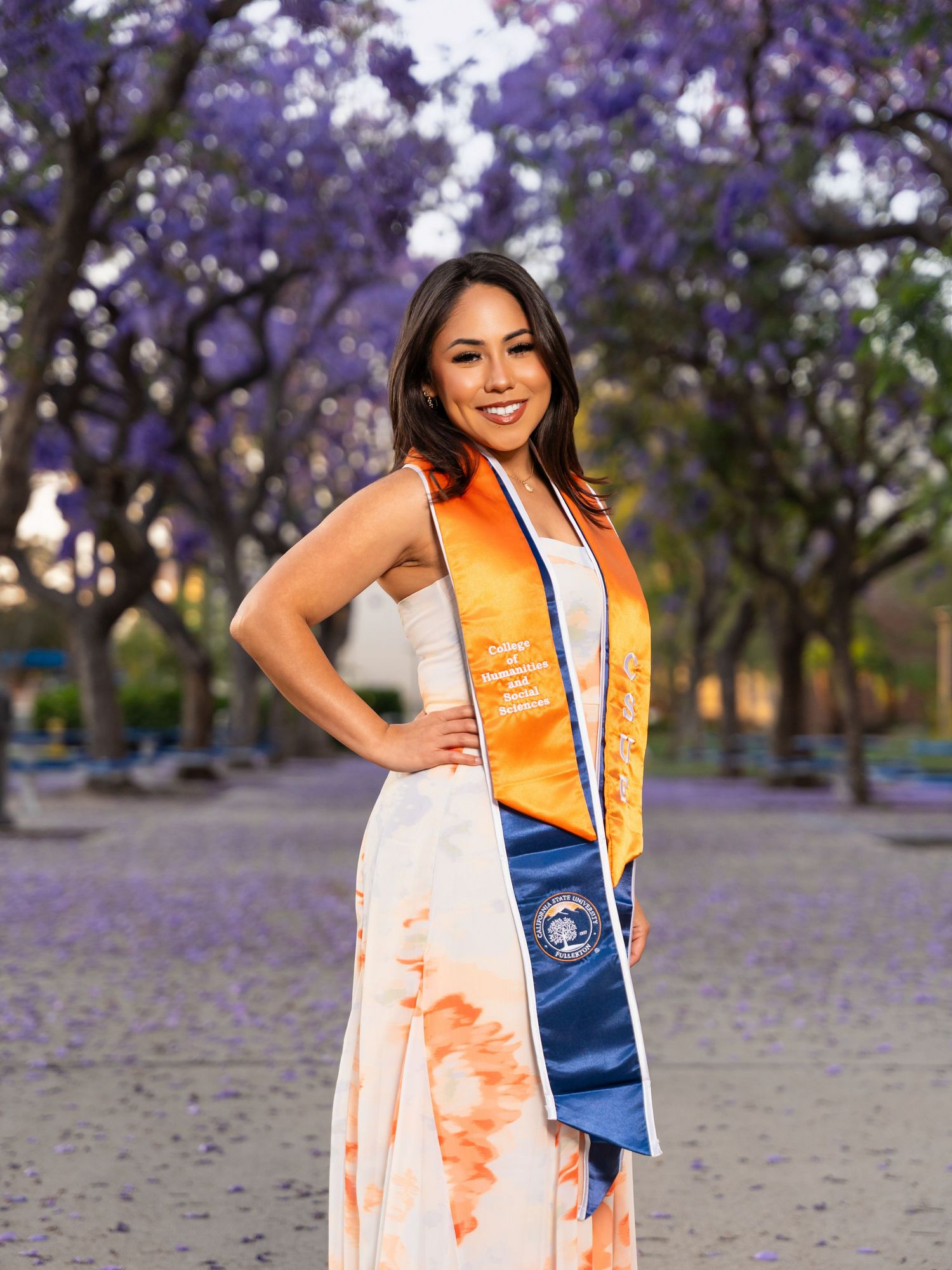 Graduate wearing orange sash and blue stole poses under blooming purple jacaranda trees during outdoor photo session.