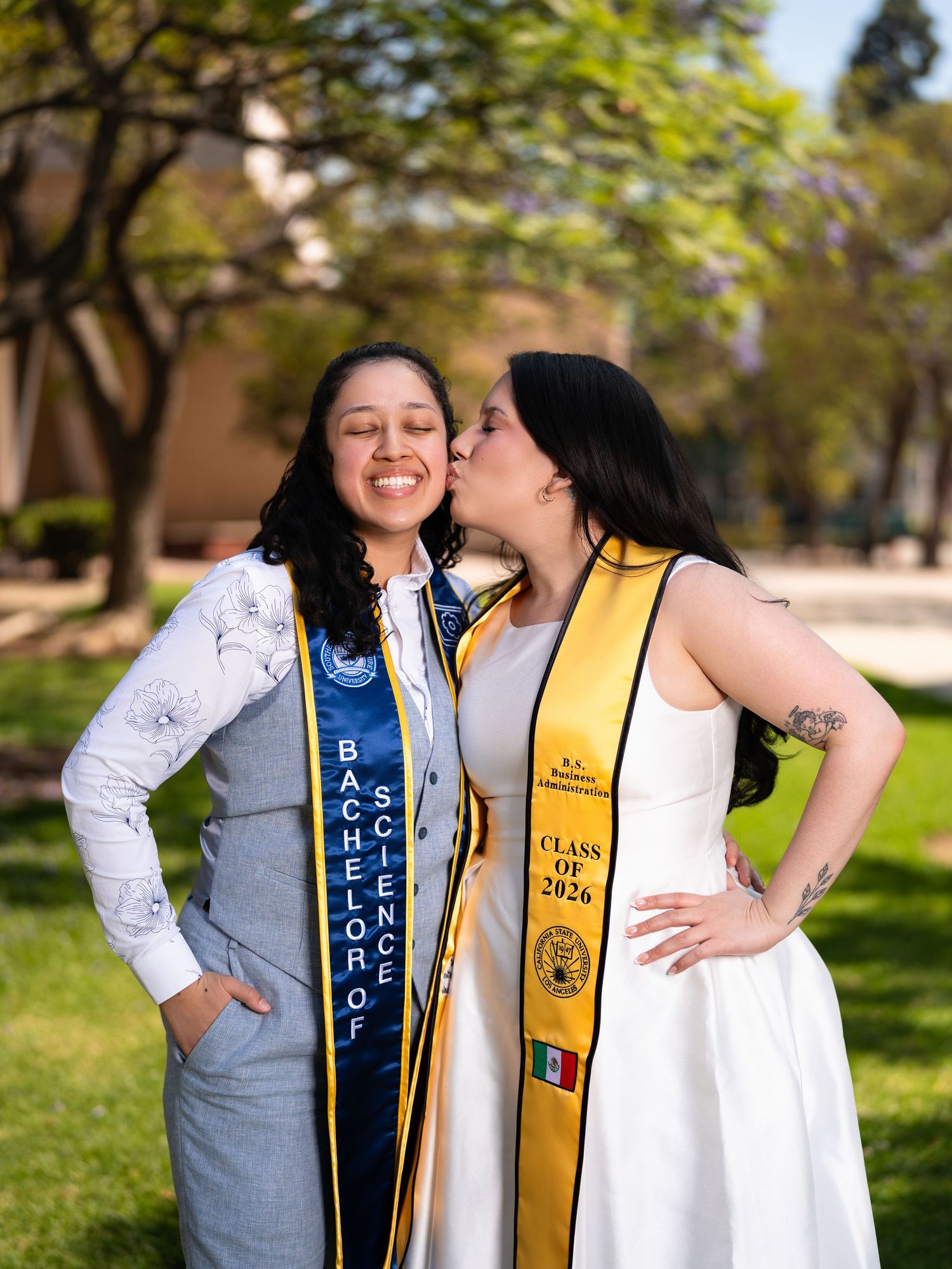 Two graduates in academic stoles, one wearing a Bachelor of Science sash, share a joyful kiss on a sunny campus lawn.