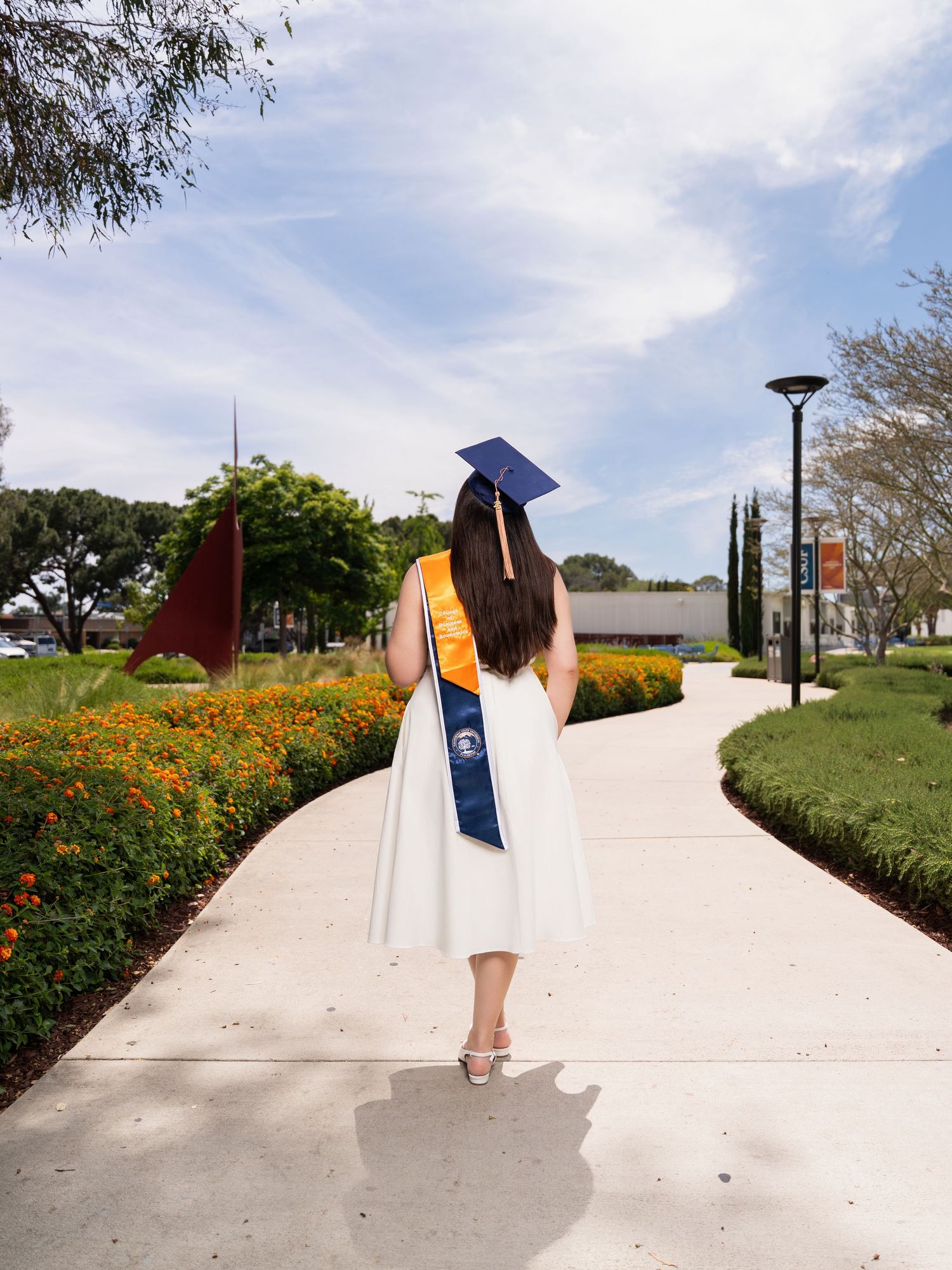 Graduate in white dress and blue sash walks along sunny campus path with orange flowers and modern sculpture.