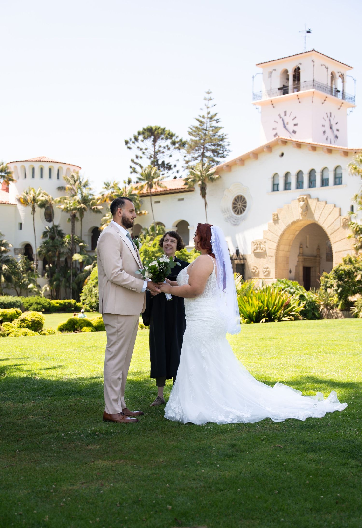 Wedding ceremony unfolding on green lawn with Spanish-style church architecture in background.