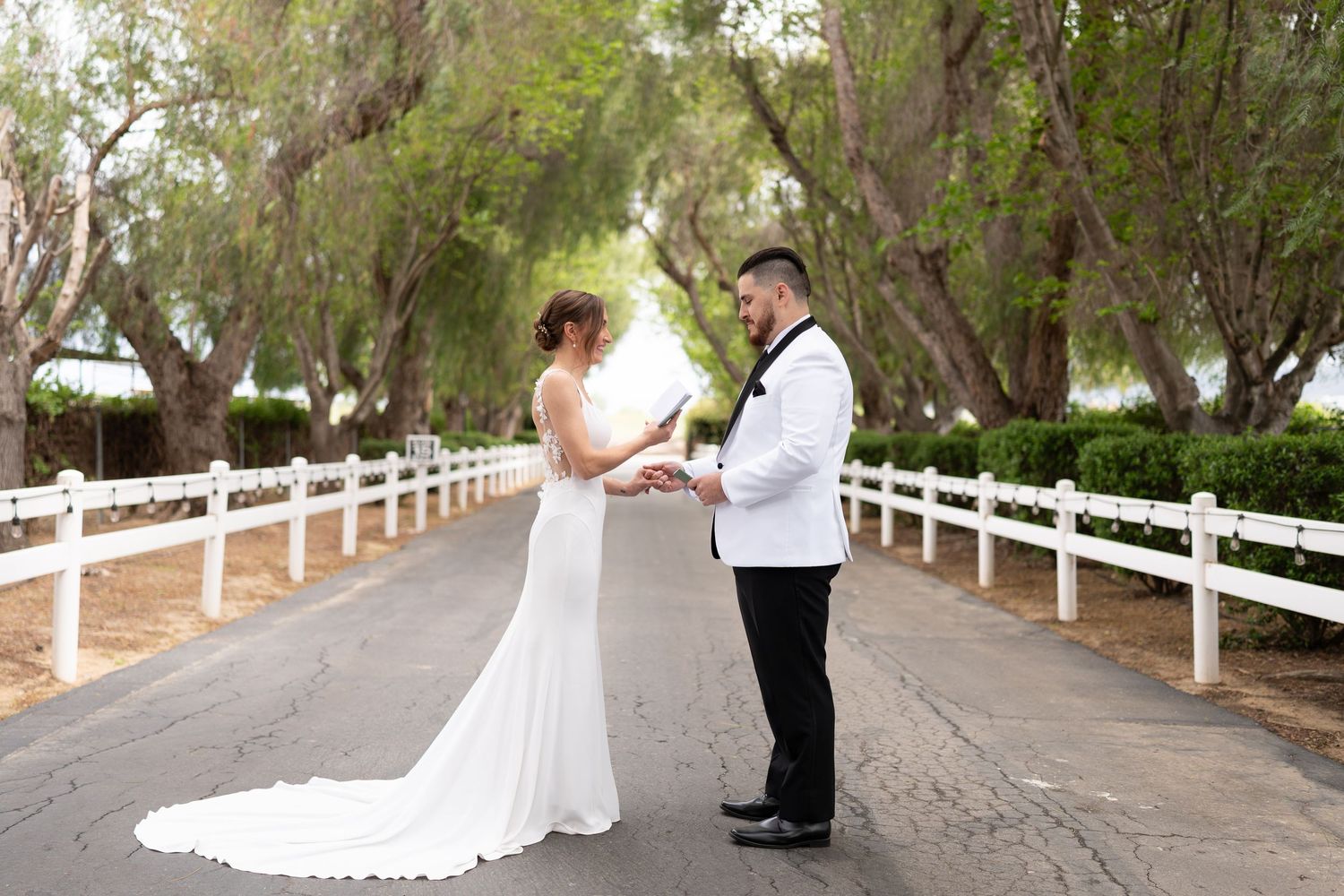 A wedding couple stands together on a tree-lined path with white fence railings.