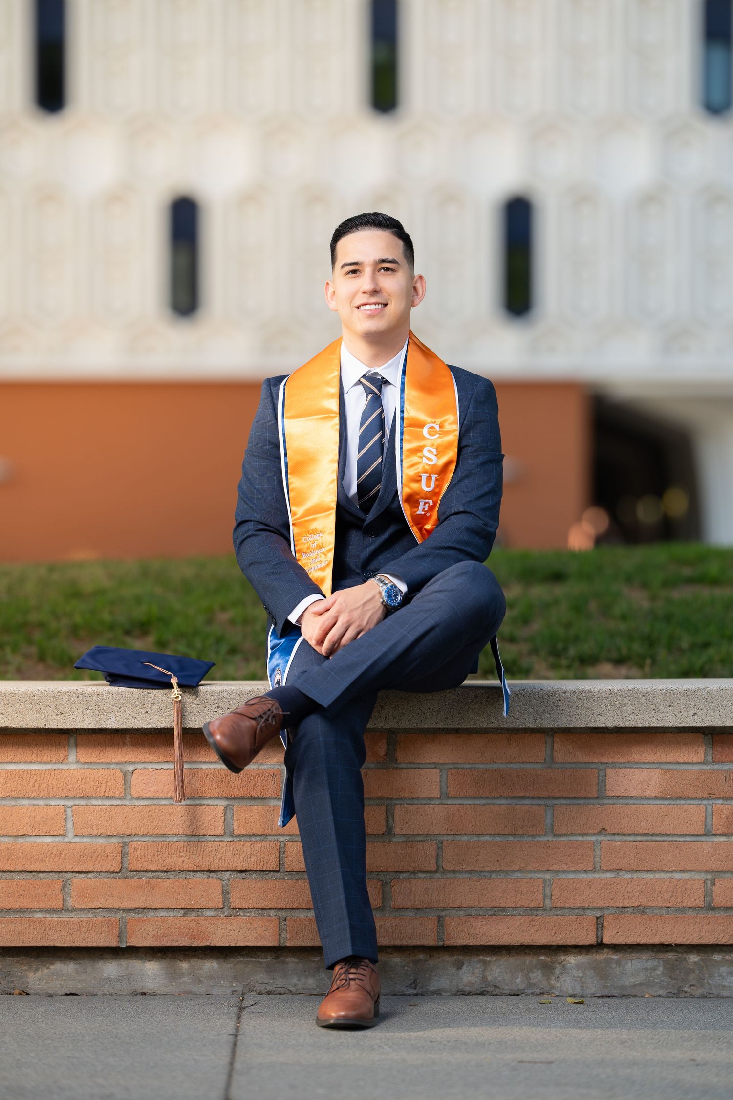 Graduate in navy suit and orange sash sits on brick ledge during outdoor graduation photo session.