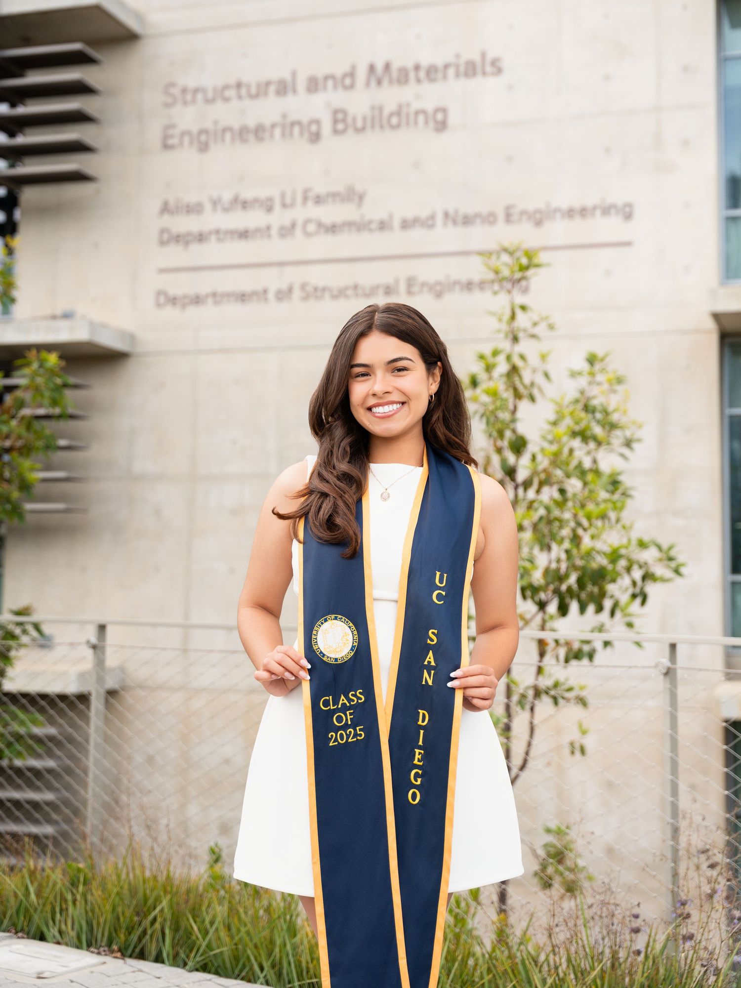 Graduate in white dress and navy blue graduation stole stands outside Structural Engineering building at university.