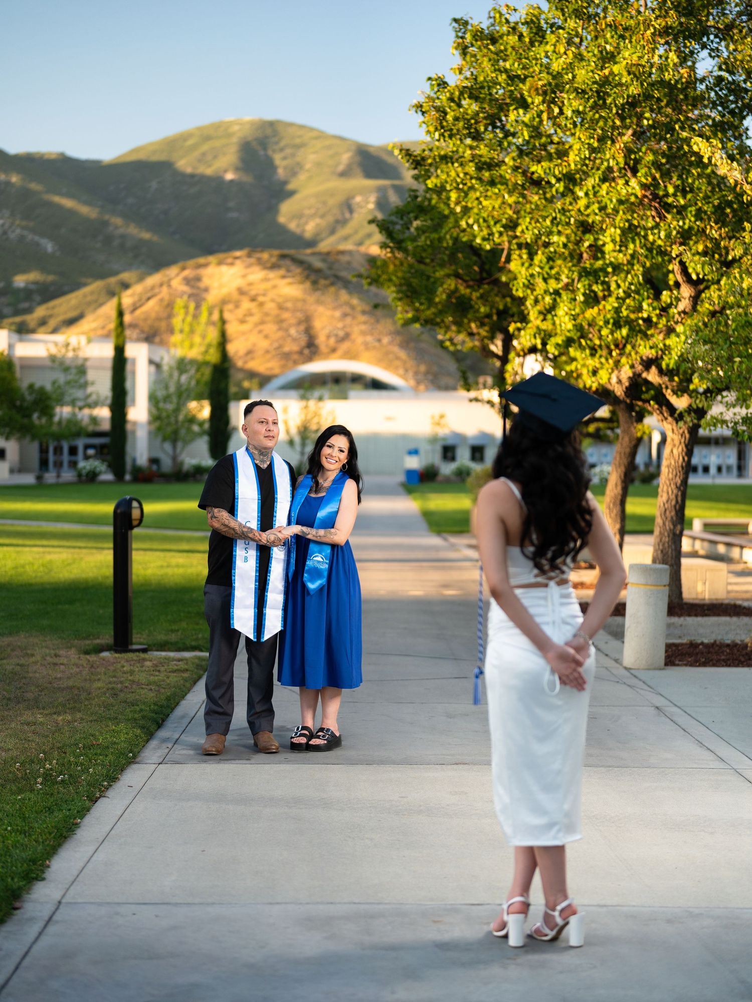 Graduate in blue cap and gown poses with family on sunny campus walkway with green hills in background.