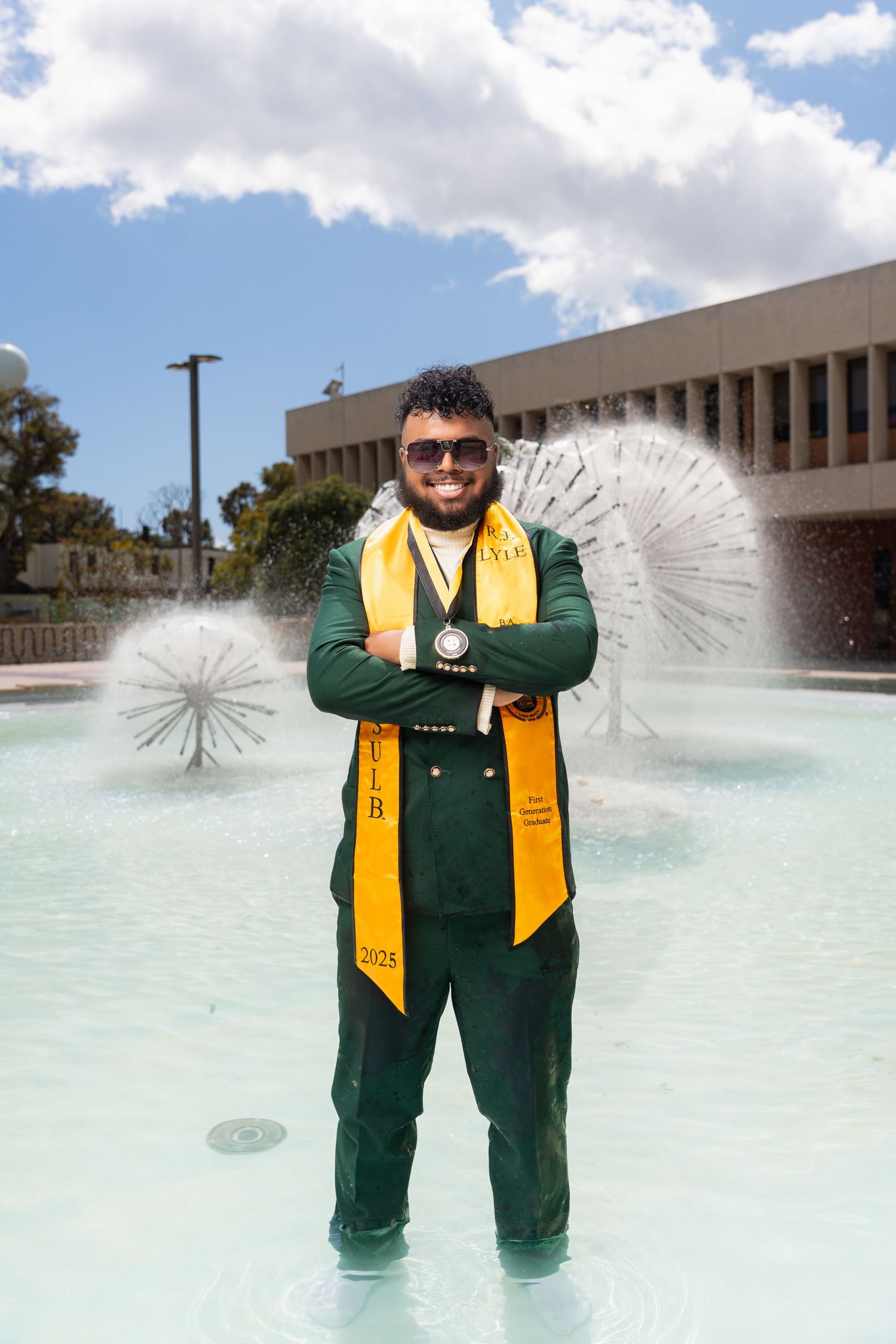 Graduate in green regalia and gold stole poses in front of a fountain during commencement celebration.