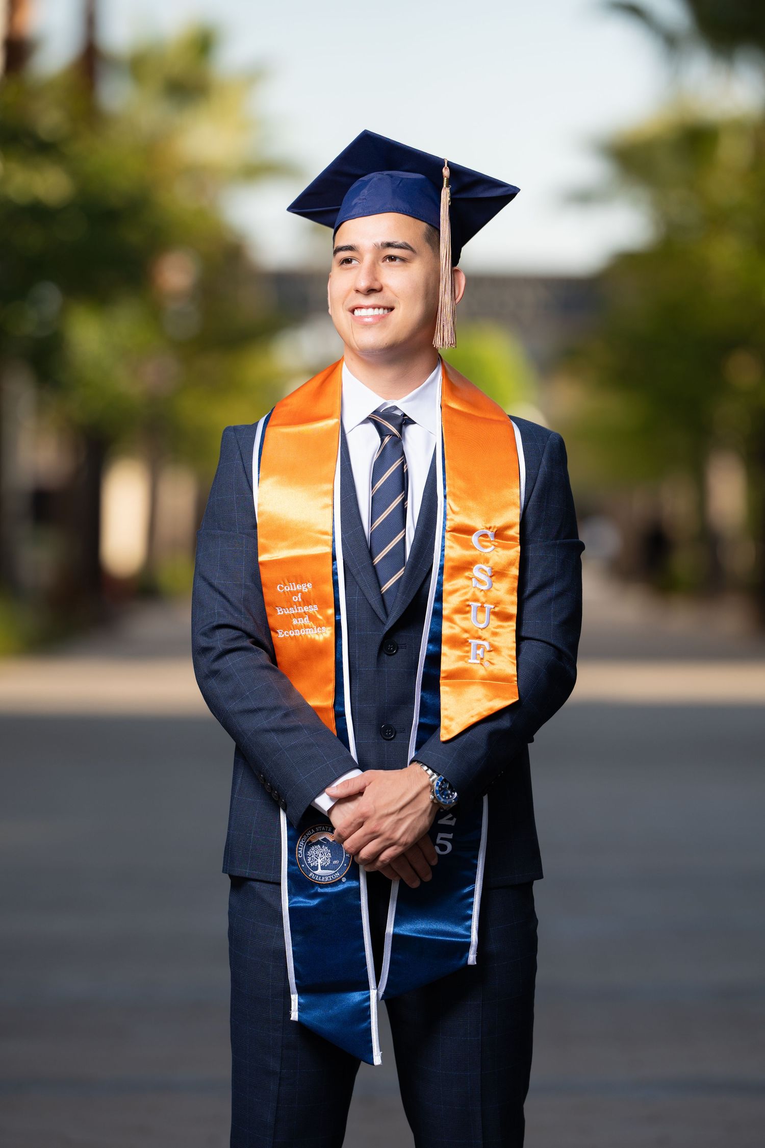 Graduate in navy suit and orange stole poses for professional graduation photos outdoors on campus.
