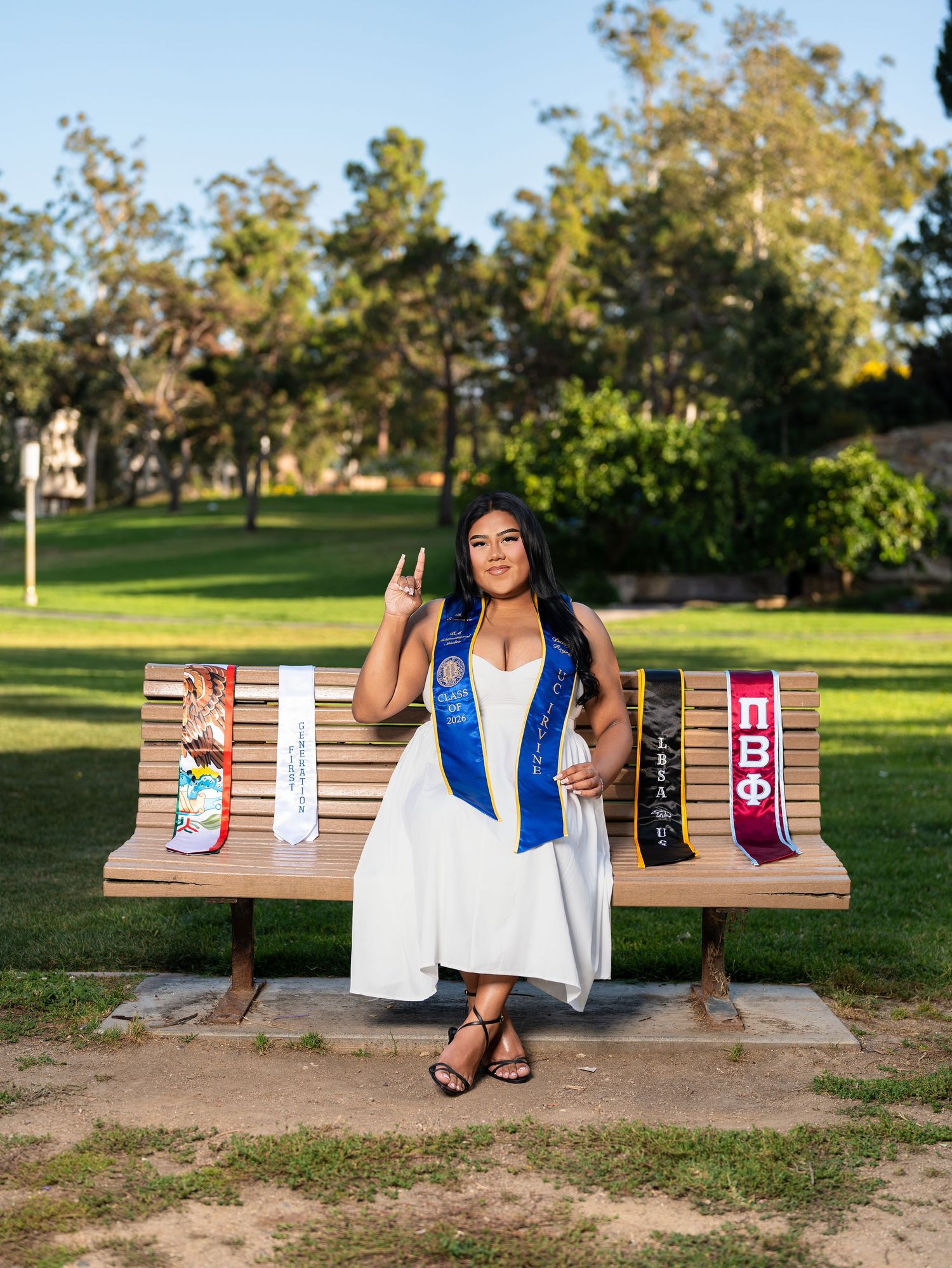 Graduate in white dress poses on bench with academic sashes and Greek letters in a sunny park setting.