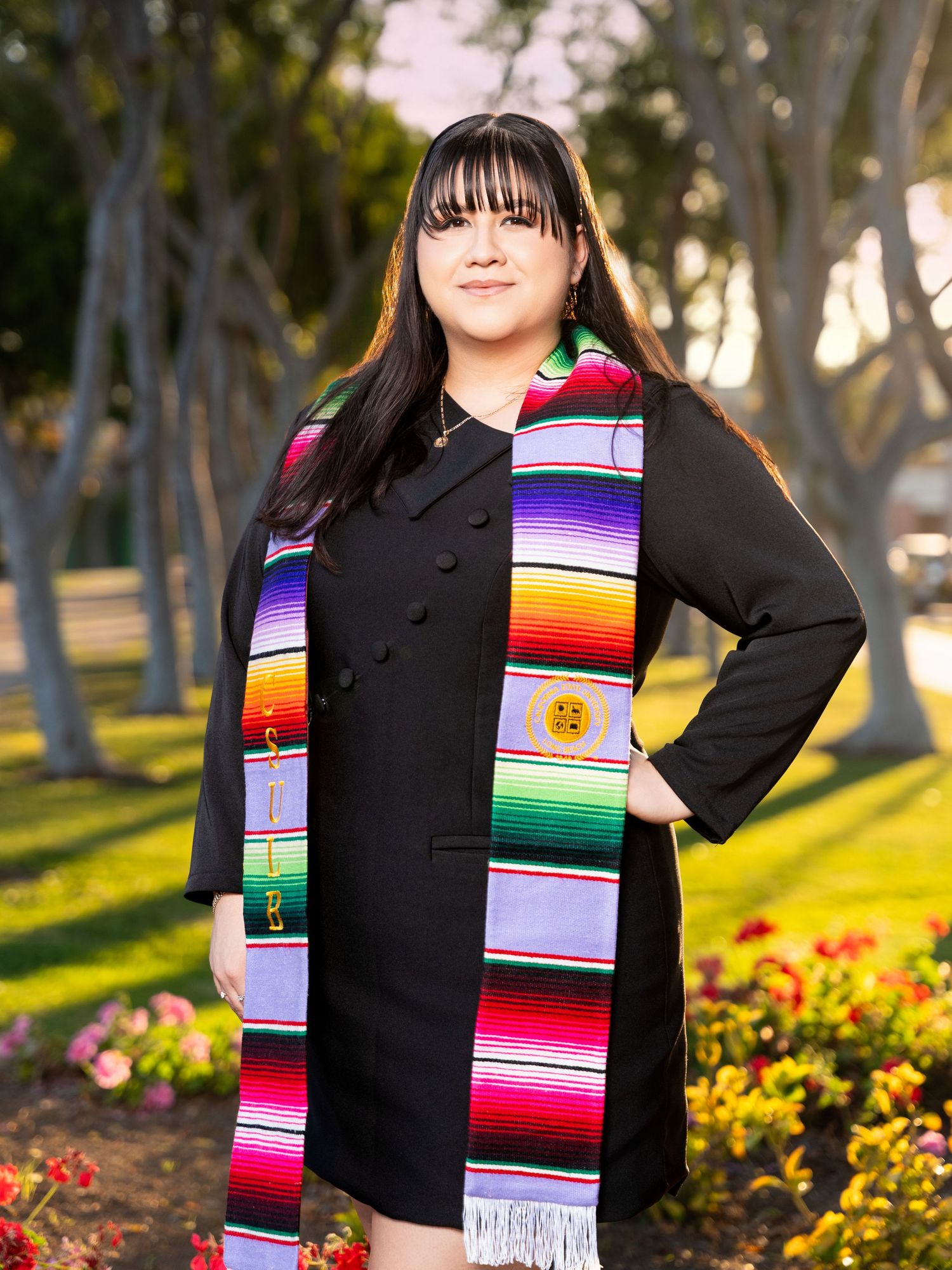 Graduate wearing black top and colorful Mexican serape graduation stole poses in outdoor garden setting at sunset.