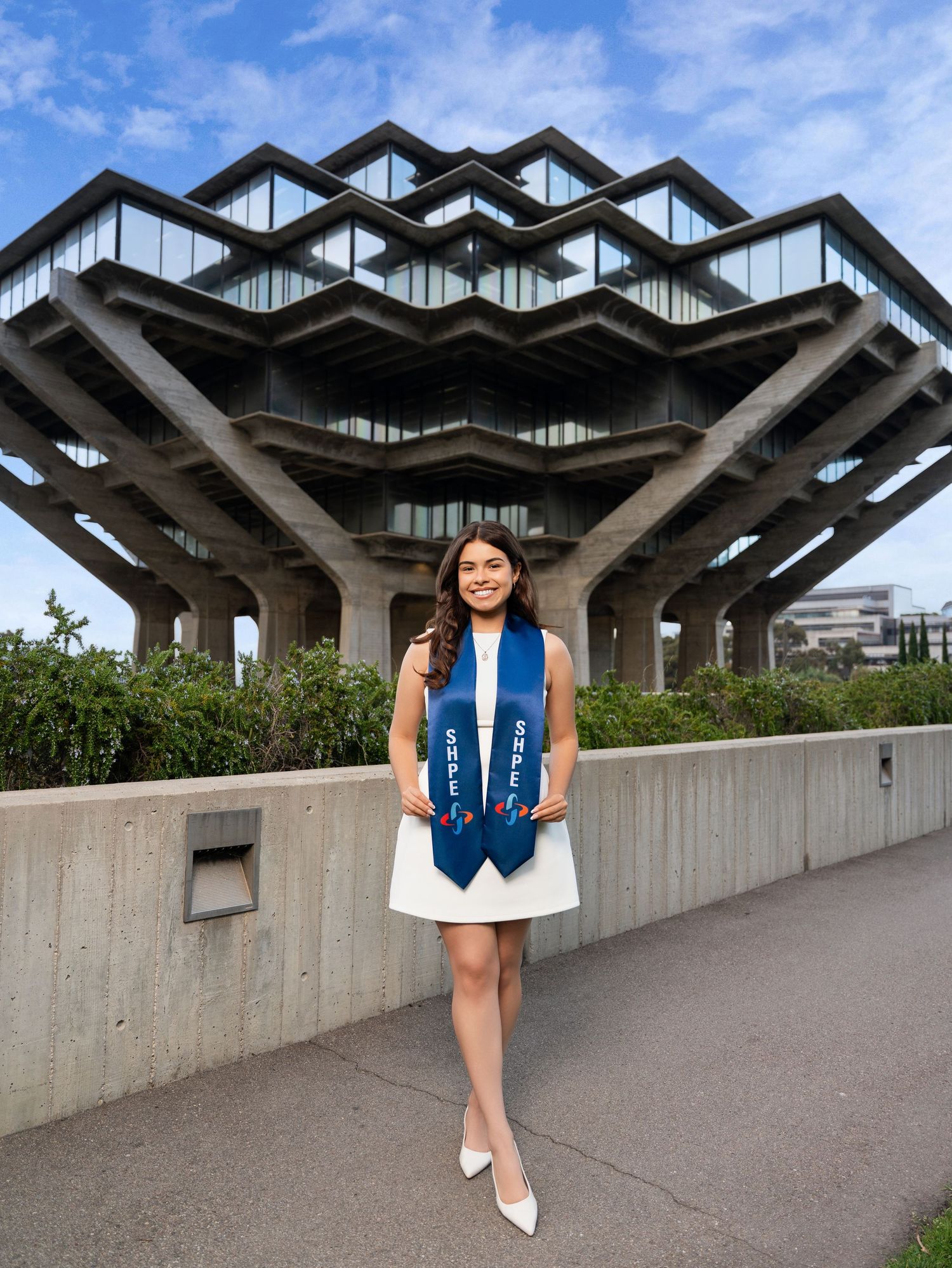 Graduate poses in front of the geometric architecture of UCSD's Geisel Library on a sunny day.