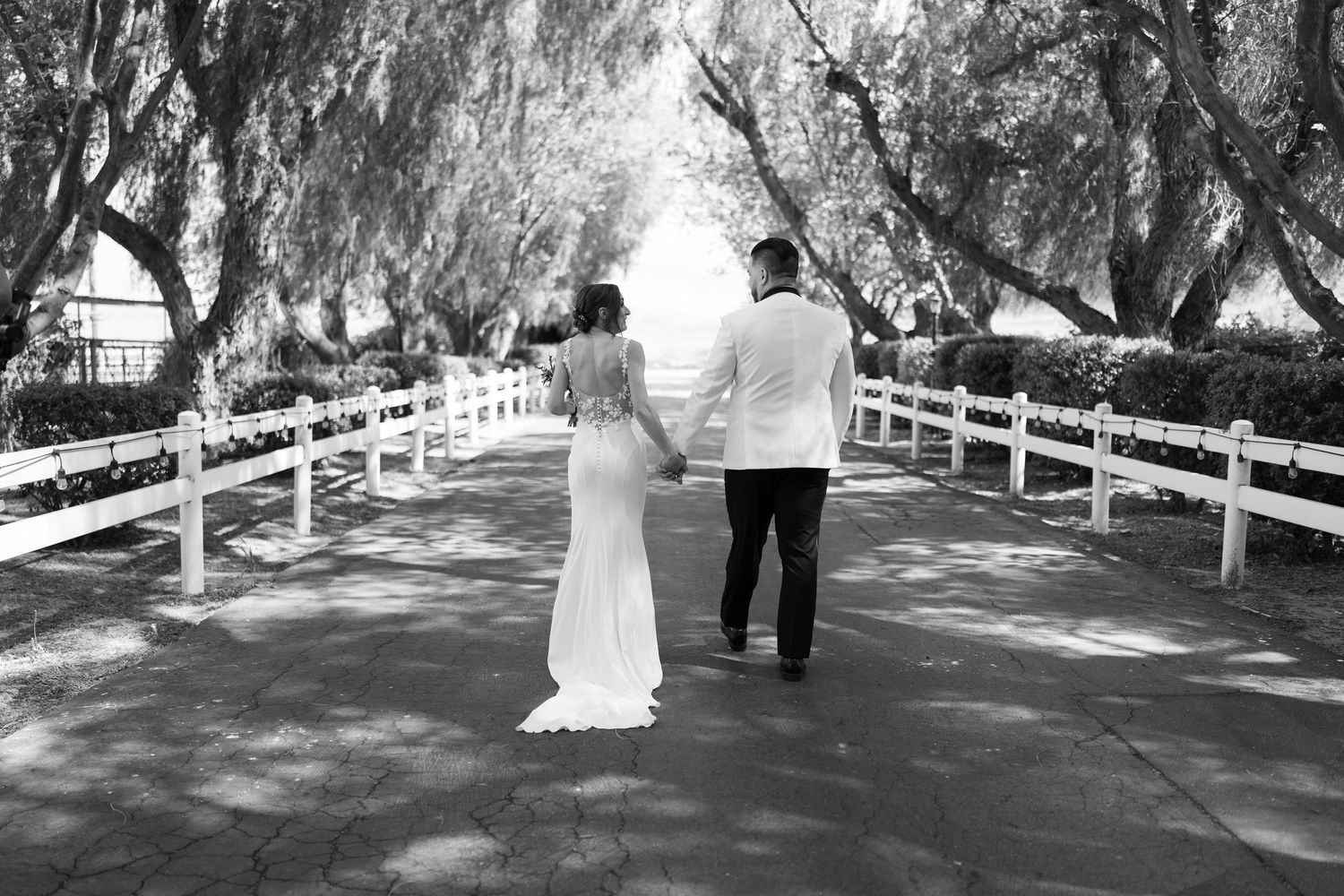 A tree-lined path with white fences leads to a romantic outdoor wedding ceremony scene.