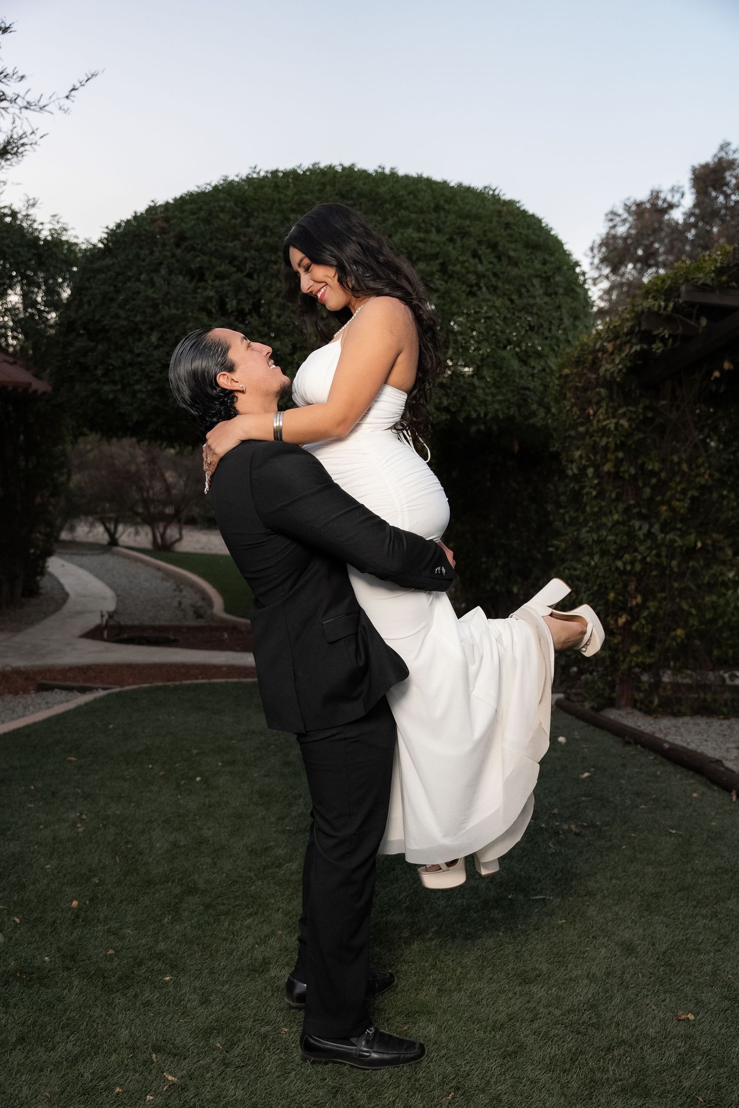 A newlywed couple performs a playful 'dip' dance move on grass with trees in the background at sunset.