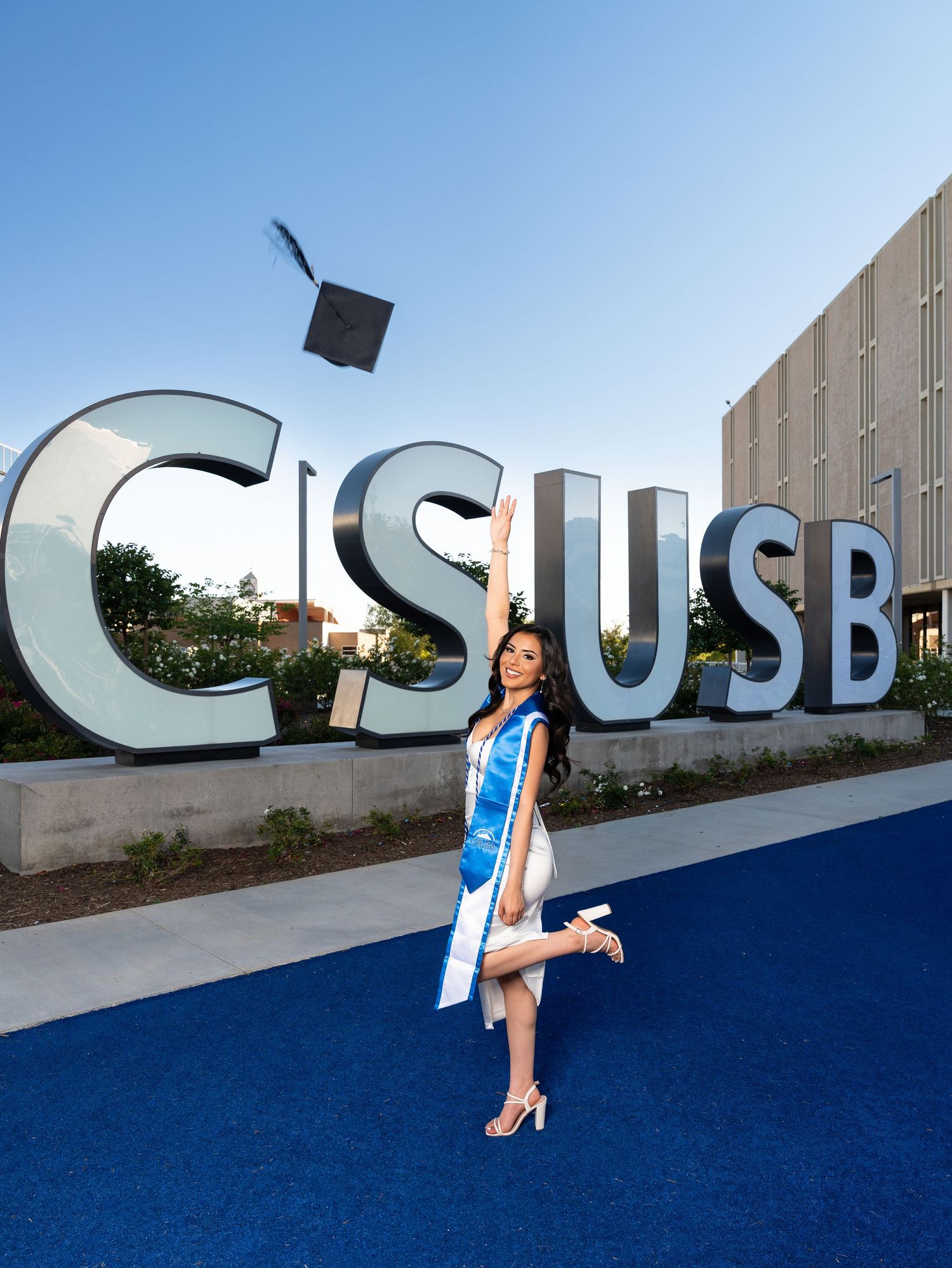 Graduate tosses cap in front of CSUSB sign, wearing white gown and blue sash on a sunny day.