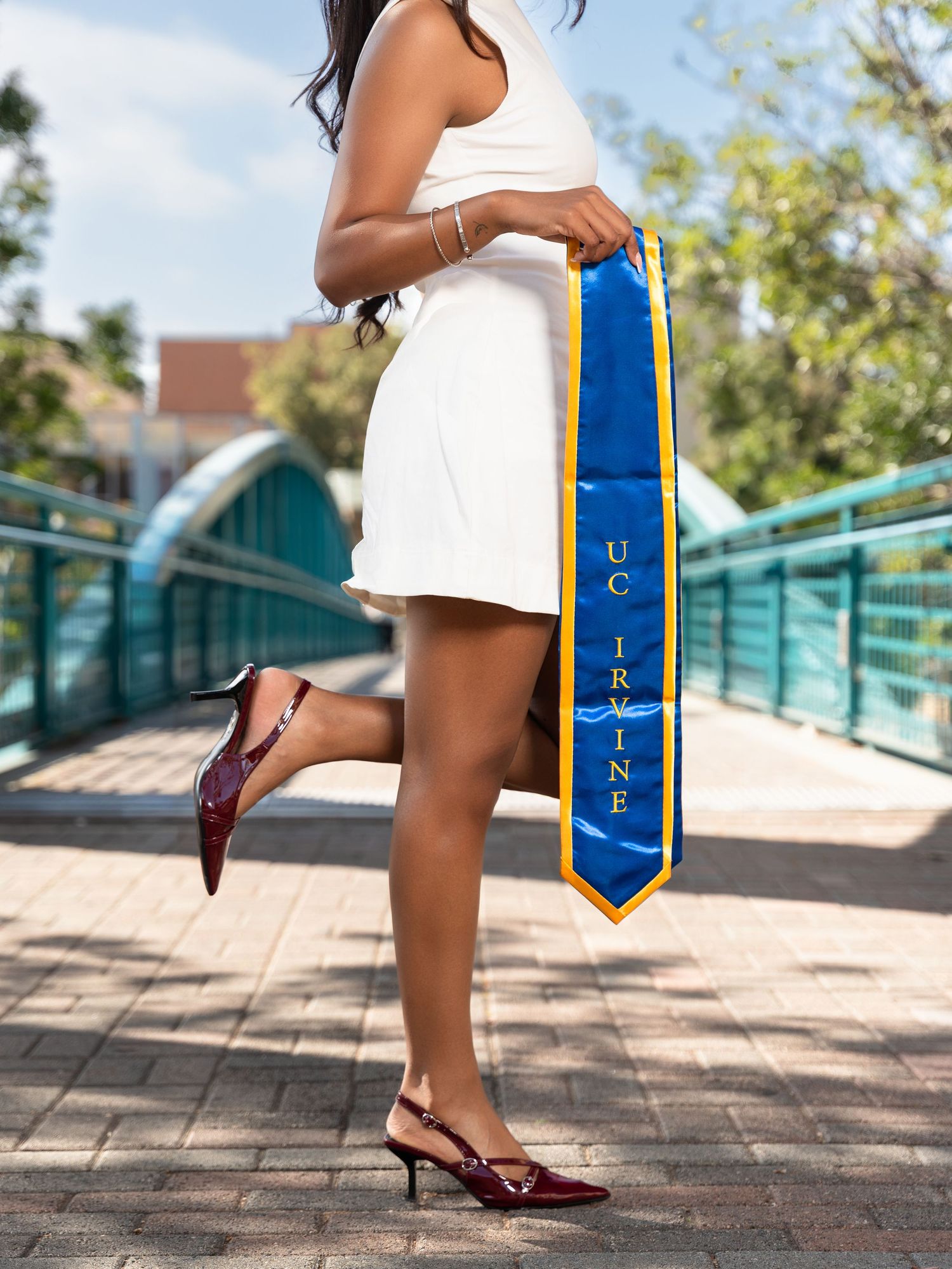 UC Irvine graduate in white dress holding blue and gold graduation stole on a bridge, wearing burgundy heels.