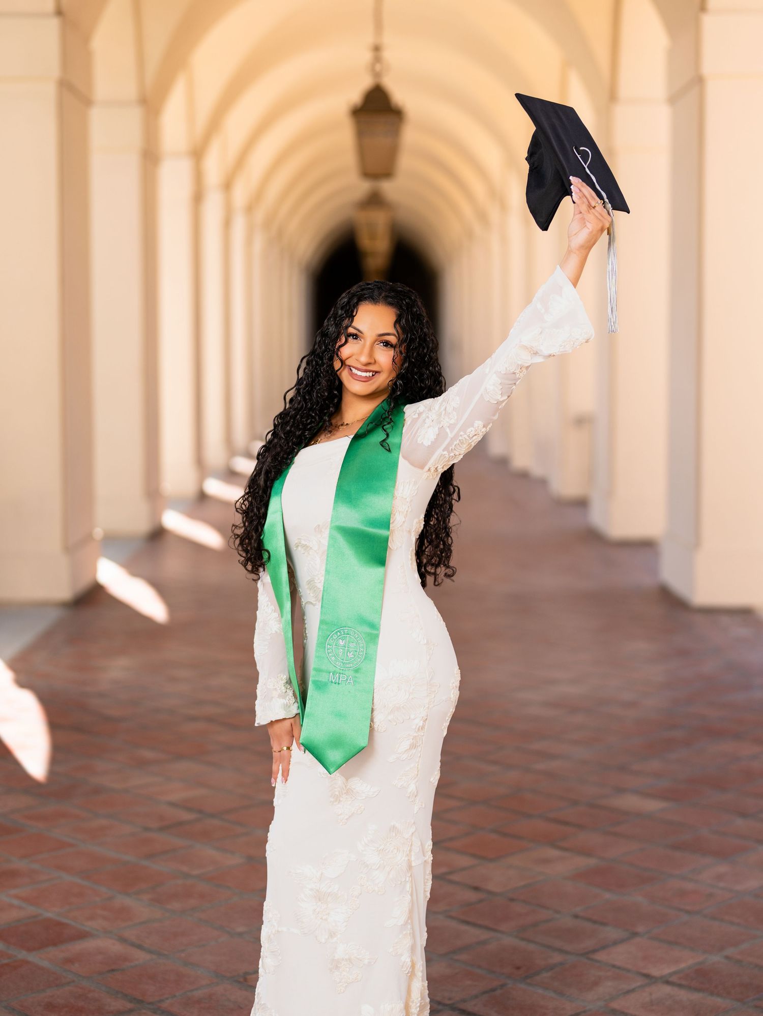 Graduate in white dress and green sash celebrates with raised cap in arched hallway during graduation photoshoot.