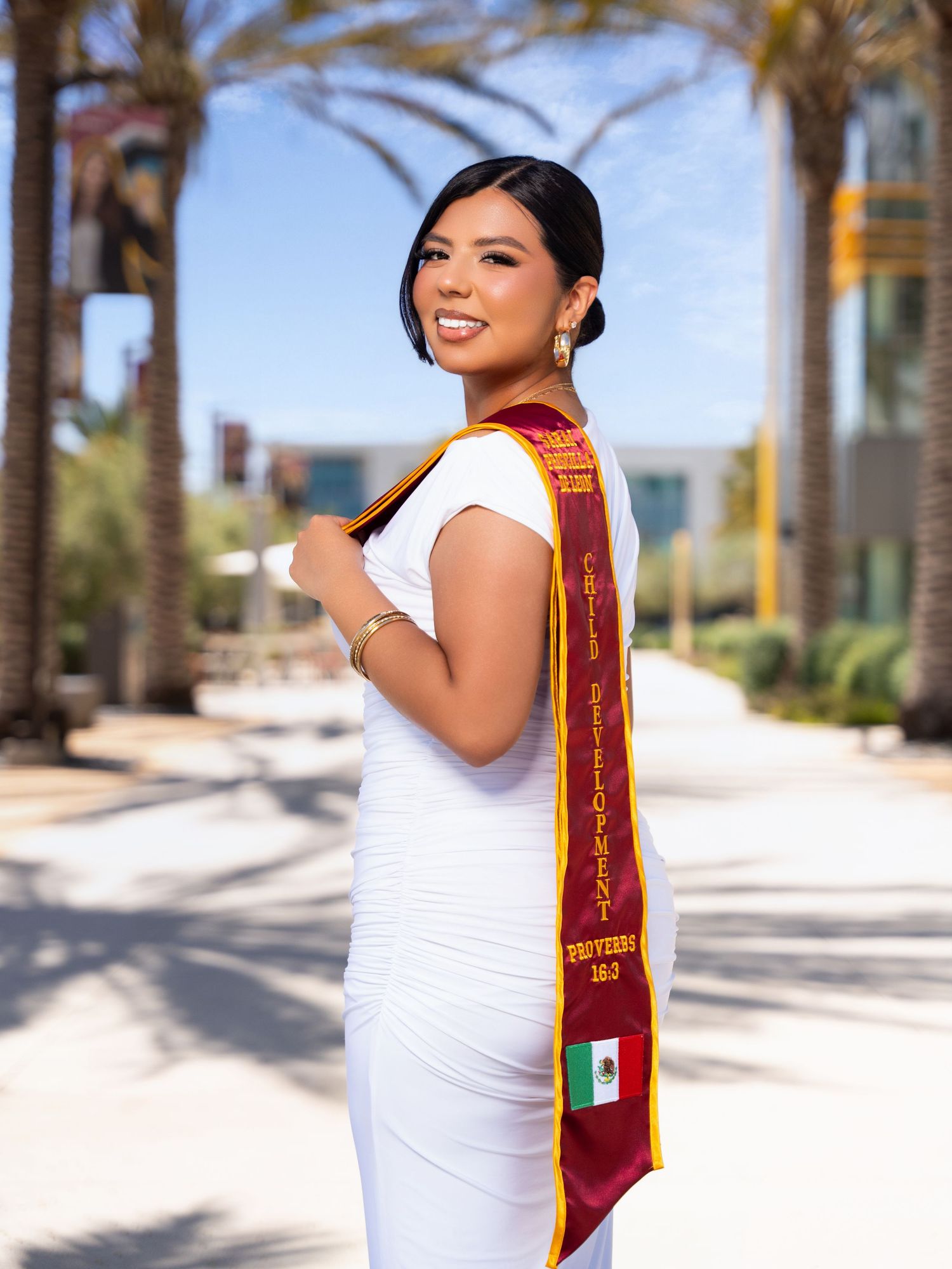Graduate in white dress and maroon graduation stole smiles while posing against palm trees on sunny day.
