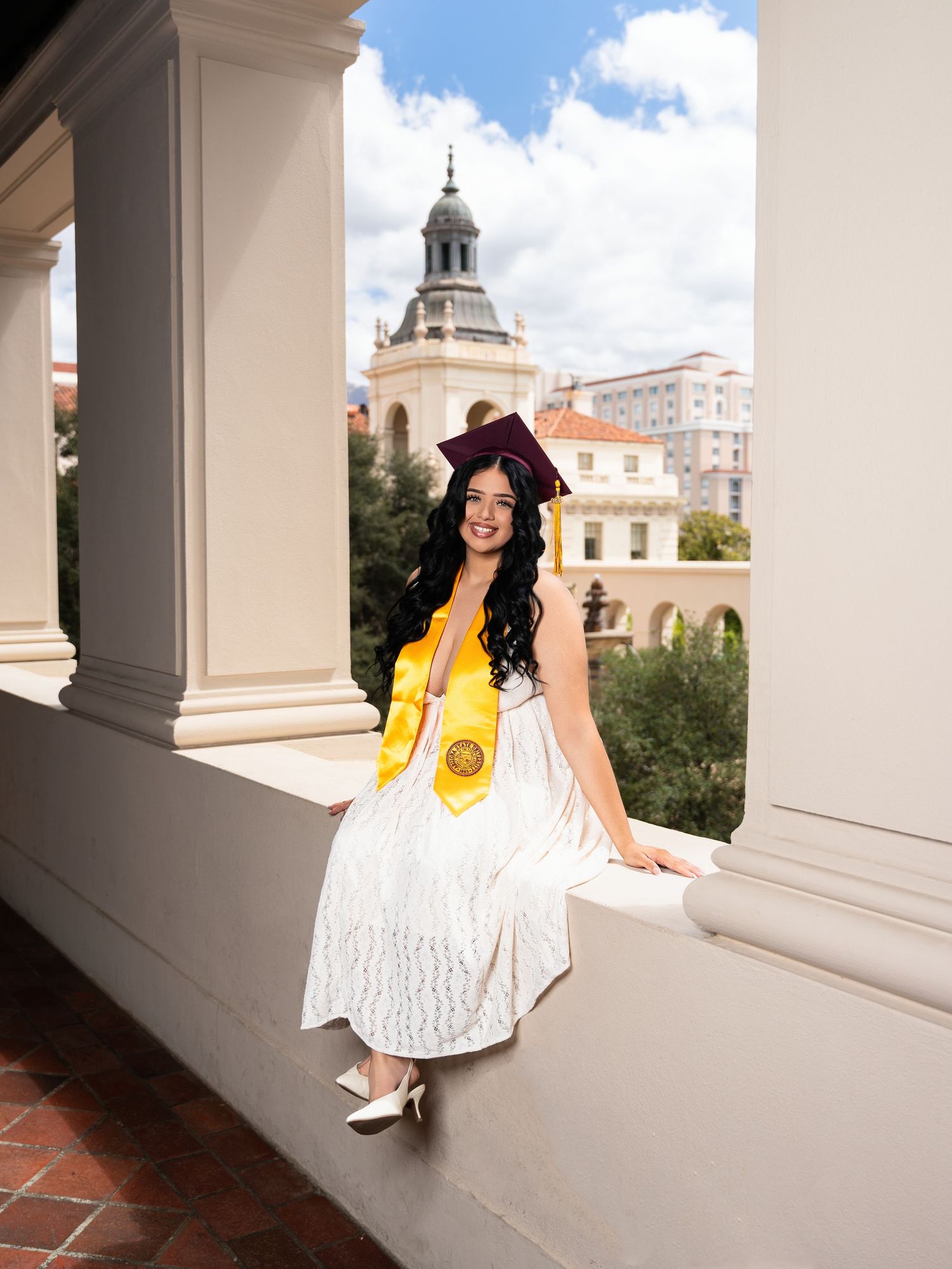 A smiling graduate in white dress, yellow sash, and maroon cap poses on a balcony with a historic building backdrop.