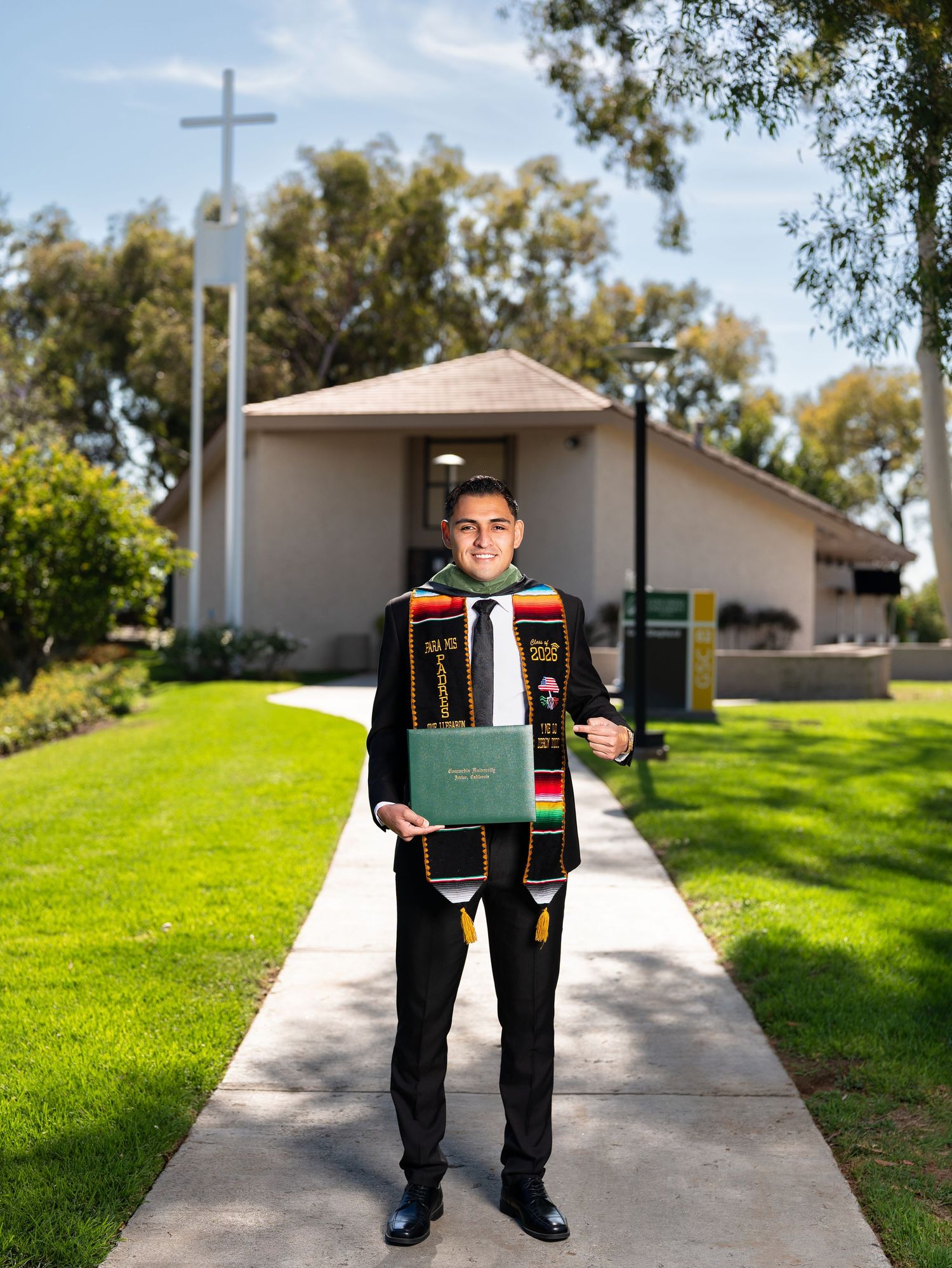 Graduate in cap and gown holding diploma outside church with cross, wearing colorful stole on sunny day.