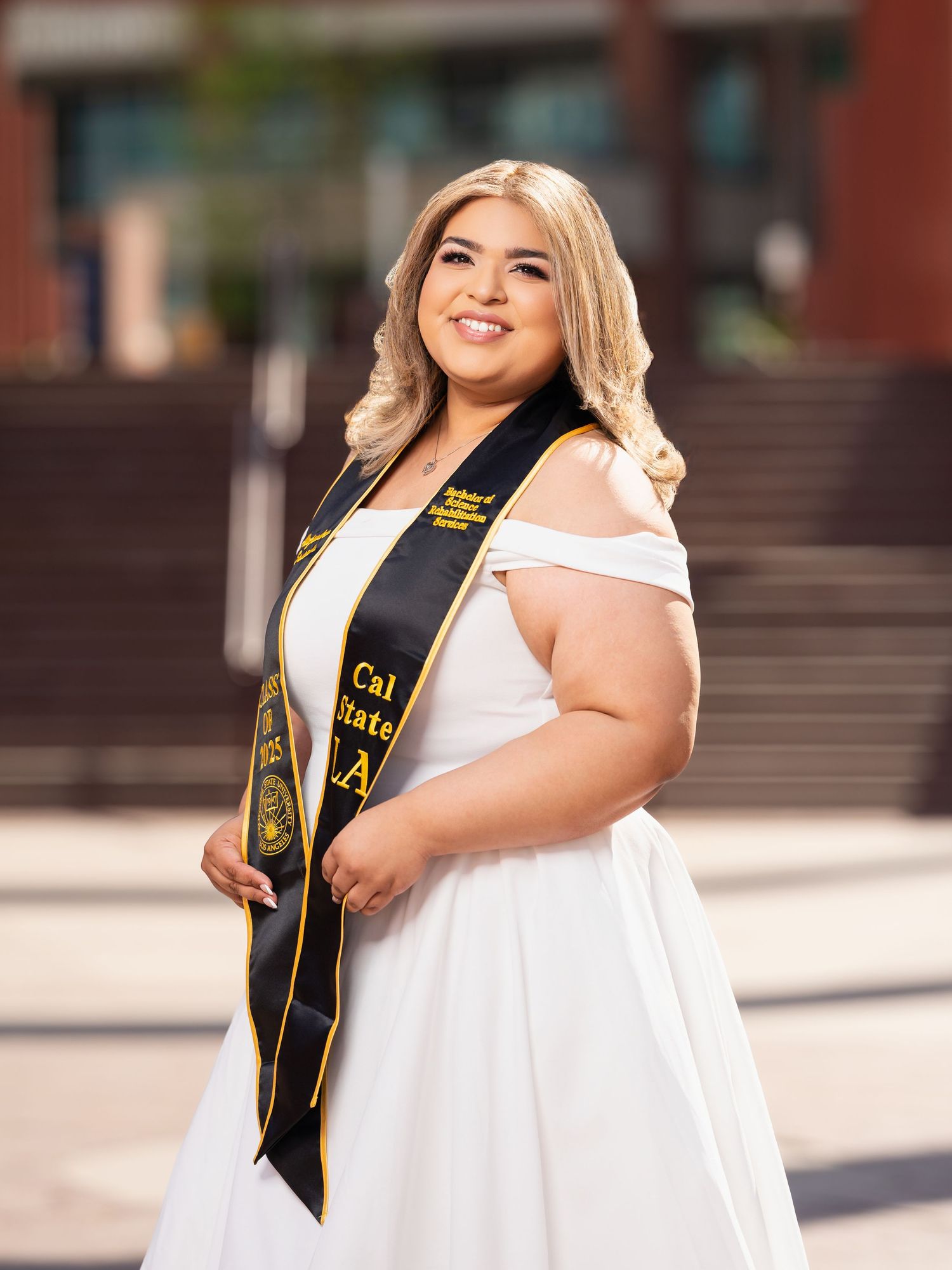 Graduate in white dress and black graduation sash poses outdoors for celebratory photos on campus.