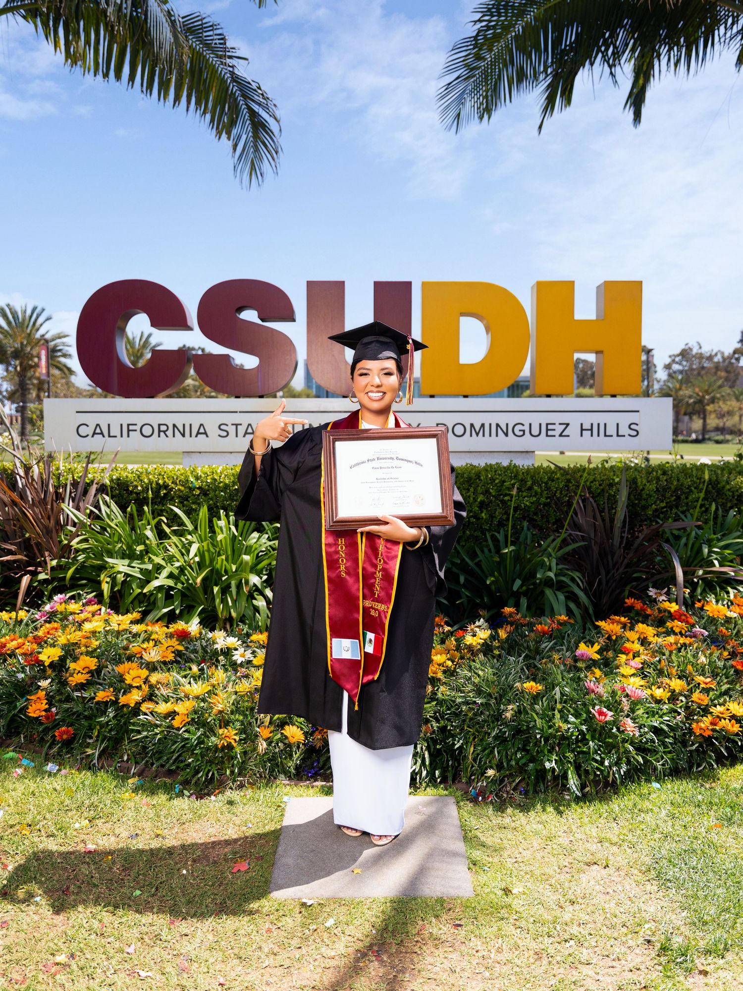 Graduate in cap and gown poses with diploma in front of CSUDH sign surrounded by palm trees and yellow flowers.