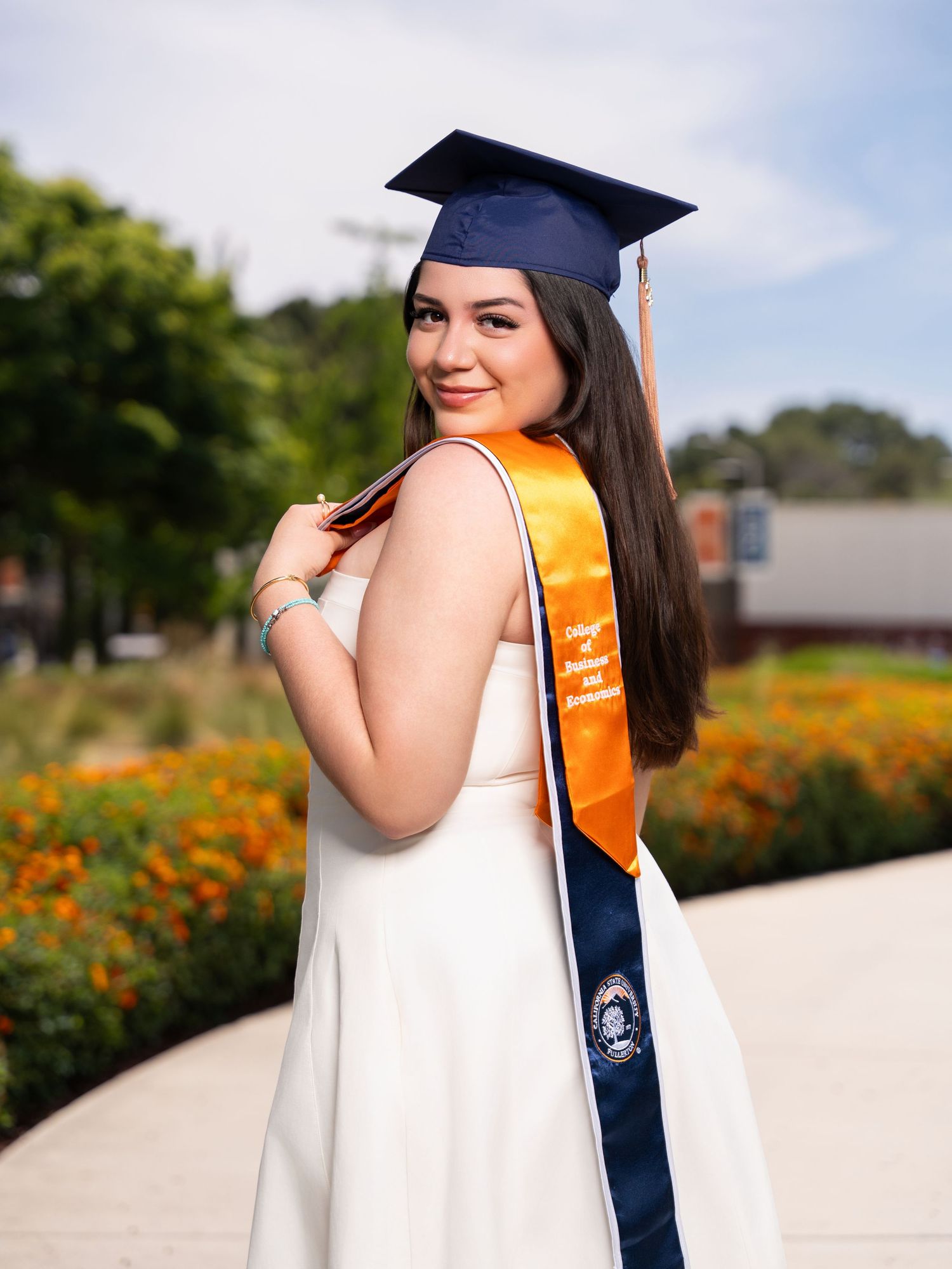 Graduate in navy cap and gown with orange sash poses outdoors for graduation photos in a garden with orange flowers.
