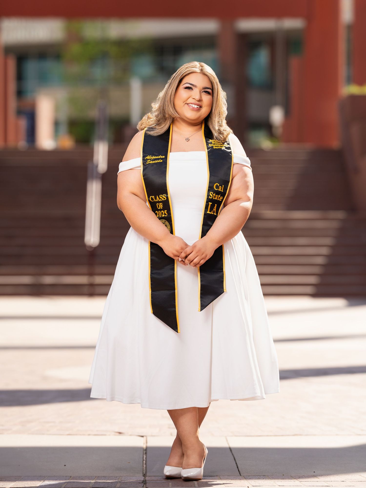 Graduate in white dress and black sash poses for celebratory photos on outdoor steps near brick building.