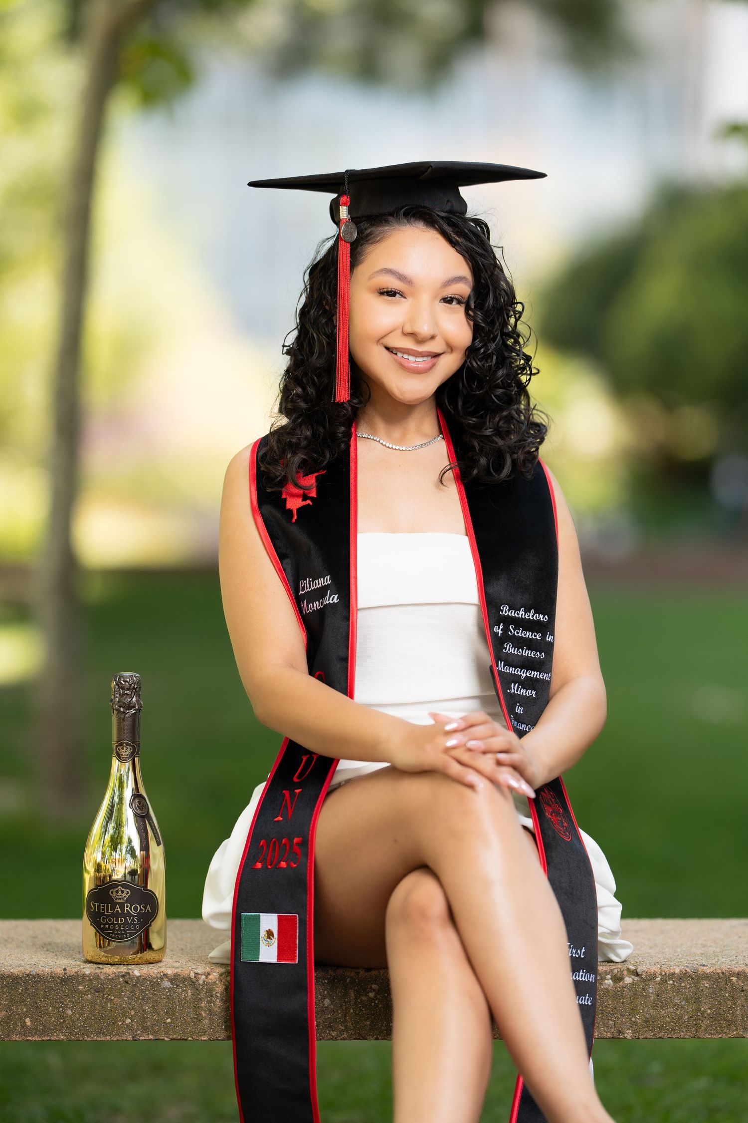Graduate in cap and gown with black and red graduation stole sits outdoors with champagne bottle for celebration photos.